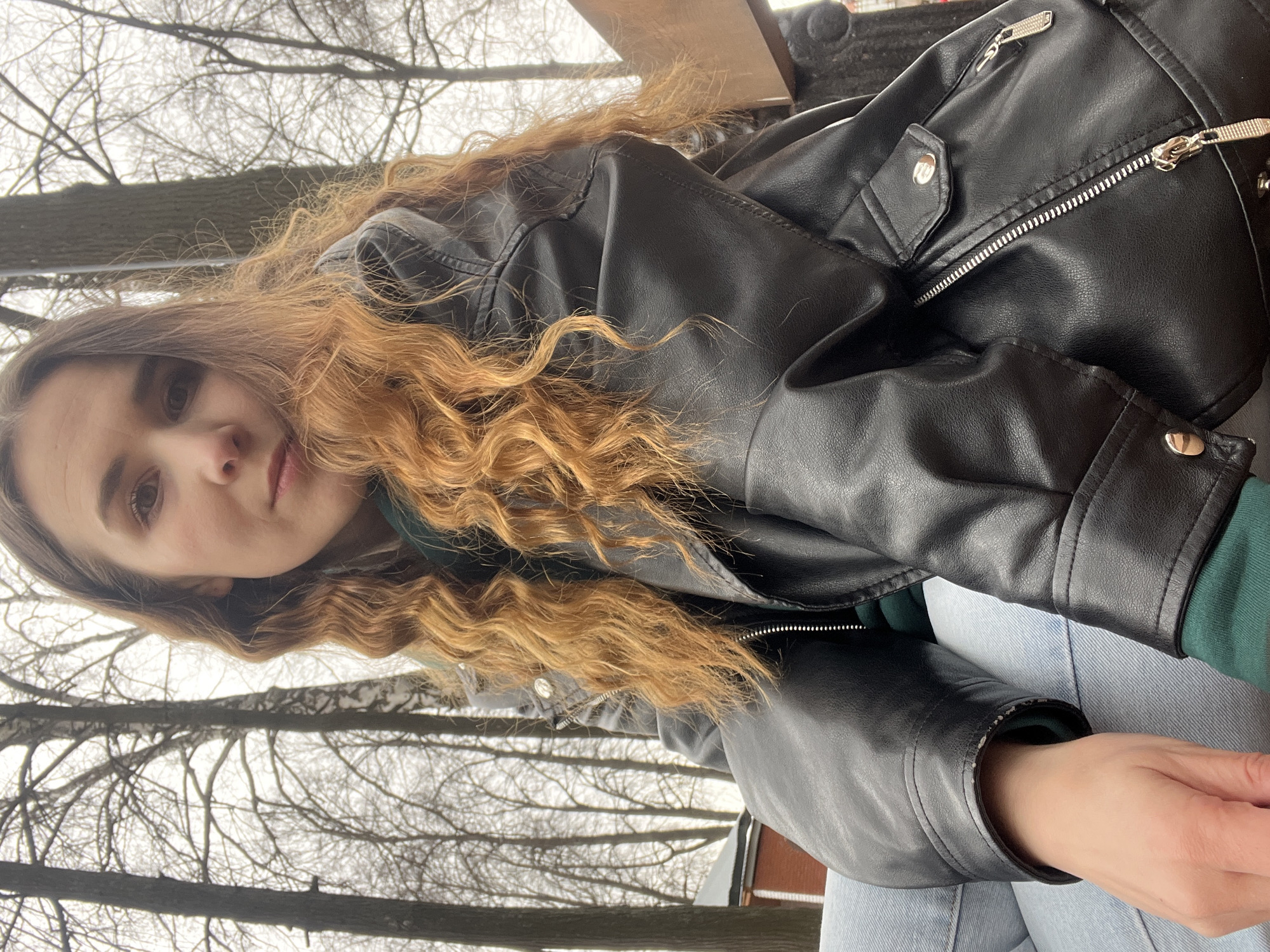 A medium, canted-angle shot depicts a woman with light skin and reddish-brown curly hair wearing a black leather jacket and light blue jeans, possibly sitting outdoors. The background shows bare trees against an overcast sky, suggesting a winter or early spring setting. No specific landmarks are visible to confirm the location in Votkinsk, Russia. The woman's expression is neutral, and her gaze is directed toward the camera.