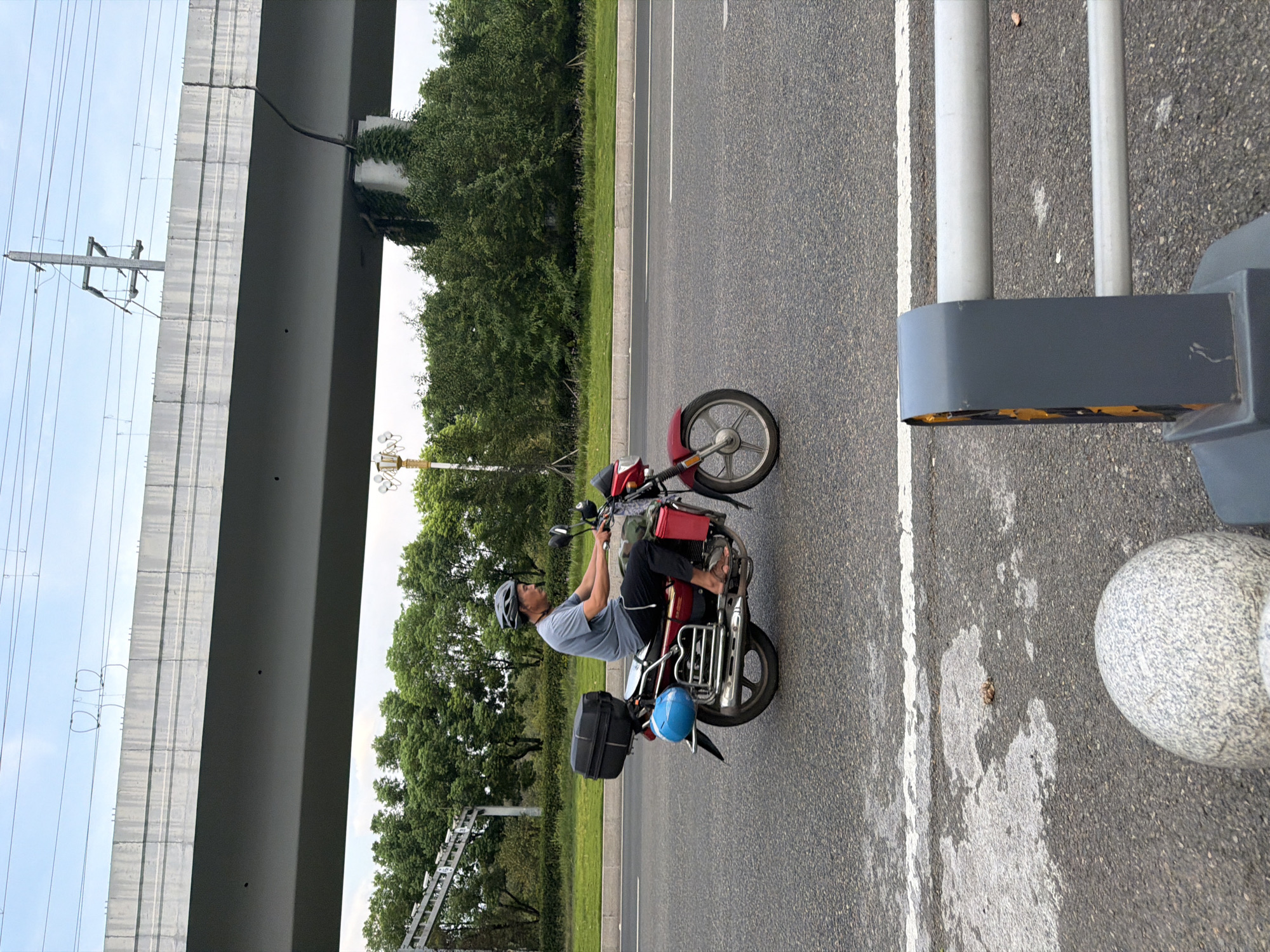 In Jiangjin, China, a man wearing a helmet is riding a red motorcycle on a paved road. The motorcycle has a black storage box on the back and a blue object on the side. The man is wearing a grey t-shirt and dark pants. In the background, there is a large concrete overpass supported by pillars, with trees and vegetation on either side of the road. Power lines are visible above the overpass. The sky is overcast with some blue patches. The foreground shows a segment of a grey railing and a large, round, speckled stone.