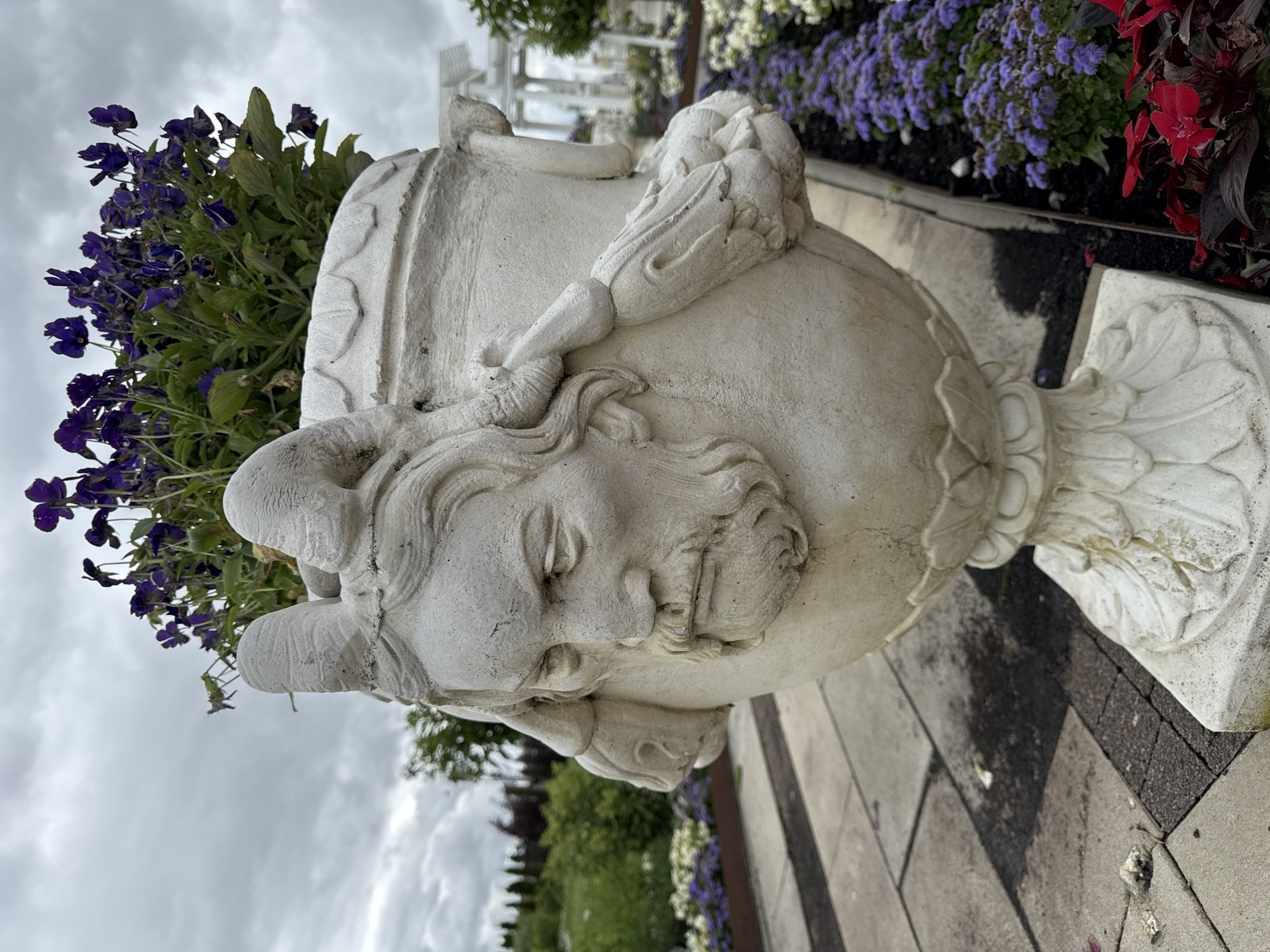 An ornately decorated, weathered stone urn, featuring a sculpted male face with a beard and horns, stands prominently in an outdoor setting. The urn is filled with vibrant purple flowers and lush green foliage. It is placed on a paved walkway with a patterned stone surface. Surrounding the urn are beds of purple and white flowers, as well as red foliage. In the background, a white pergola-like structure and trees are visible, suggesting a garden or park environment. The sky is overcast with clouds. The location is identified as Bruzdeilynas, Lithuania.