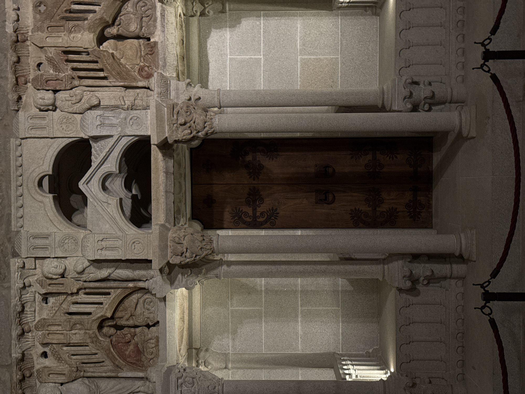This is a medium shot, eye-level, full-frame, interior photograph of an ornate stone doorway with a wooden door, located in the city of Santiago De Compostela, Spain. The doorway is framed by carved stone pillars and an elaborate stone archway above. The wooden door is dark brown and features decorative metal hardware, including scrollwork on the hinges and handles. Flanking the door are two large, fluted stone columns topped with carved capitals depicting foliage and possibly animal figures. The wall behind the columns is constructed of large, square granite blocks. The floor is also made of stone. In the foreground, a red rope is strung between two black stanchions, indicating a boundary. The scene is dimly lit, creating shadows that emphasize the textures of the stone carvings.