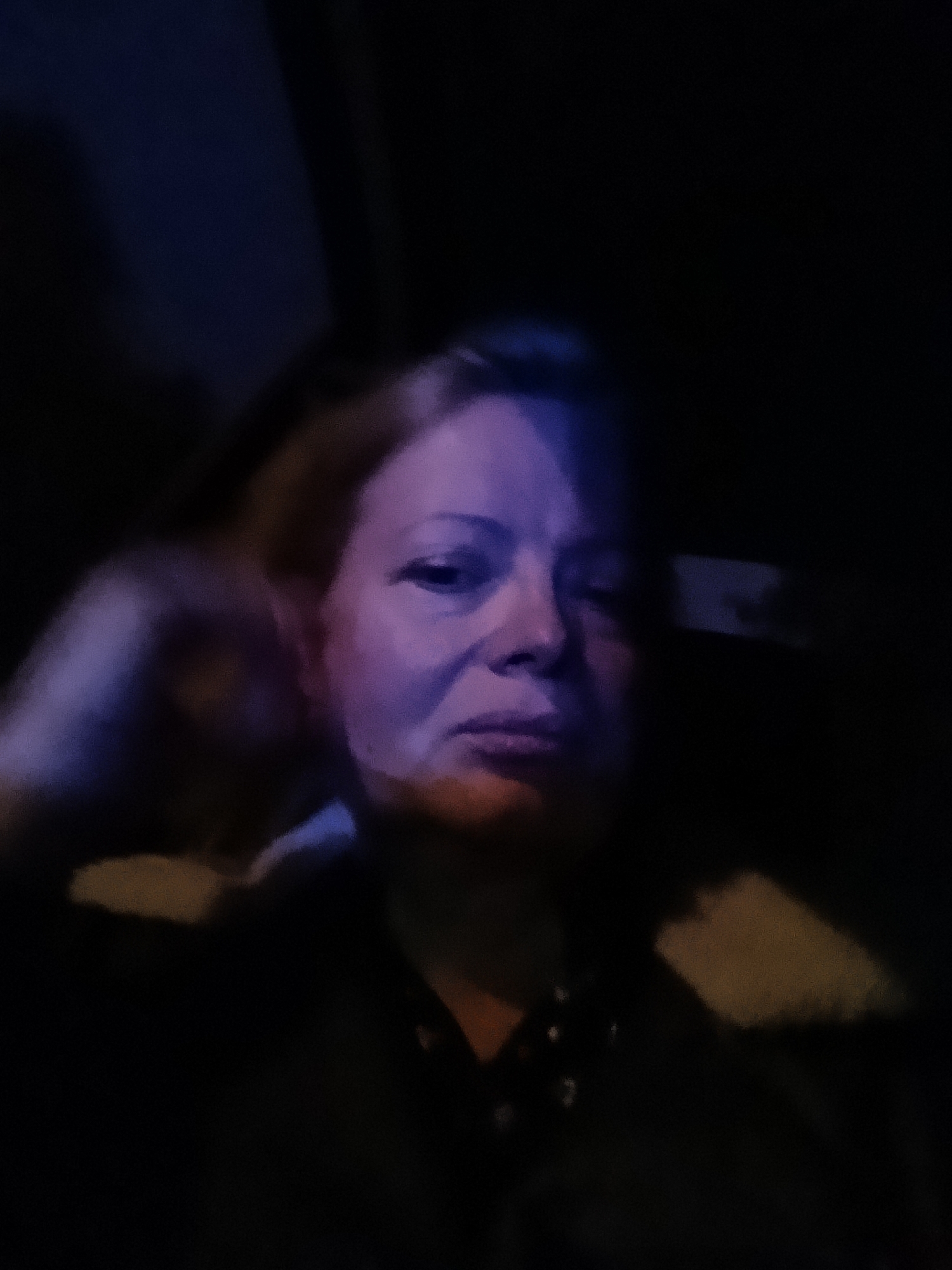 A portrait photograph captures an adult woman in a dimly lit, almost dark environment. Her face is the central focus, illuminated predominantly by a cool, blue-purple light that casts over her right cheek and forehead, creating a striking contrast with the surrounding darkness. A warmer, faint glow is also visible on her chin. Her expression appears calm, perhaps slightly serious or contemplative. She has light-colored hair, visible around her face, and is wearing dark clothing, possibly a collared top. The background is indistinct and deeply shadowed, offering no discernible details or objects. The overall impression suggests either an indoor setting at night or an extremely dark exterior, with artificial light sources providing the primary illumination. The image quality is somewhat grainy, consistent with low-light conditions. No specific activity is depicted, and no text is visible. The scene could be set in Chaikovsky, Russia.