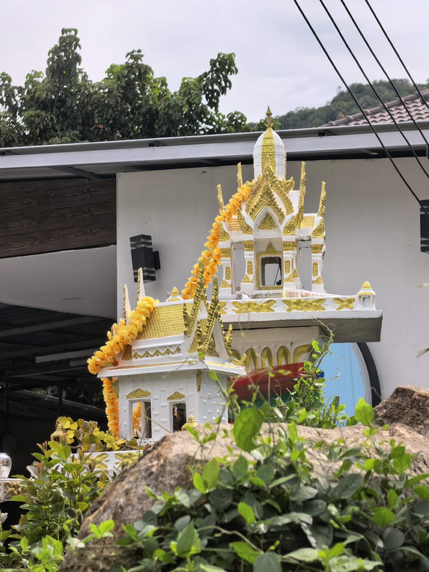 This is a medium-shot, eye-level outdoor photograph taken in Ko Samui District, Thailand. The main subject is a miniature shrine, elaborately decorated with white and gold accents, and adorned with a garland of bright yellow flowers. The shrine sits atop a ledge or base, partially obscured by lush green foliage and a large rock in the foreground. Behind the shrine, a white building wall with black light fixtures is visible. Power lines traverse the upper portion of the frame, against a slightly overcast sky. The background shows a residential building with a tiled roof and trees on a hillside, suggesting a semi-urban or suburban setting. No people are visible in the frame.