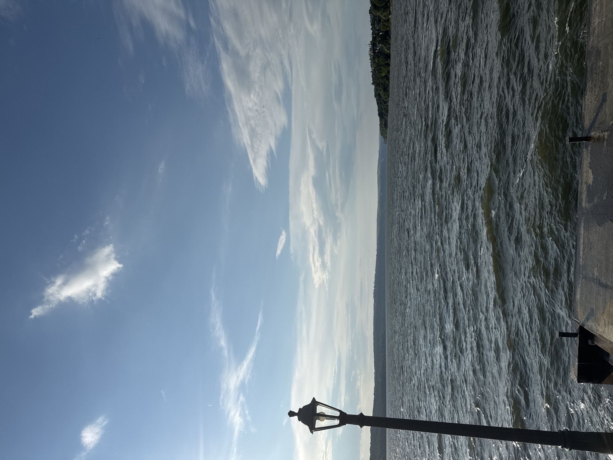 The image shows a body of water with moderate waves under a bright blue sky with scattered white clouds. In the foreground, on the left, is a dark-colored lamppost. A concrete pier or dock is visible in the lower portion of the image. The background features a distant, tree-covered shoreline with some structures visible. The scene appears to be a waterfront area, possibly a park or promenade, with the lamppost suggesting a man-made element within a natural setting. The provided location of Korzhevo, Moldova, places this scene within a specific geographical context.