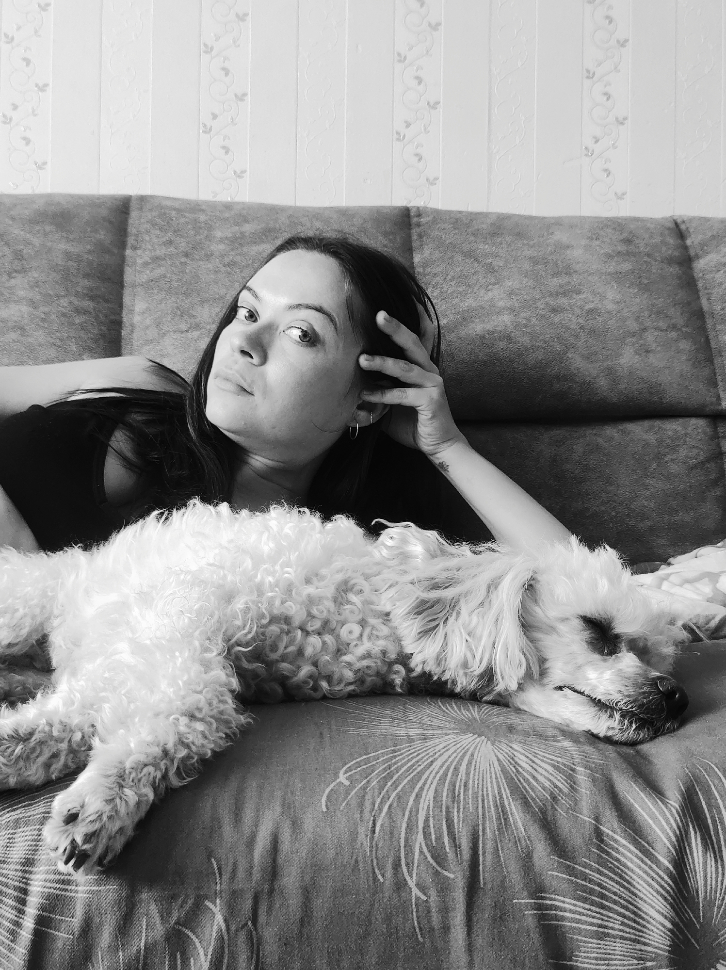 A woman lies on a patterned sofa next to a sleeping dog. The scene is in black and white. The woman rests her head in her hand and looks towards the camera. The dog is a light-colored, curly-haired breed. The background is a wall with a subtle, vertical pattern. The location is Chaikovsky, Russia.