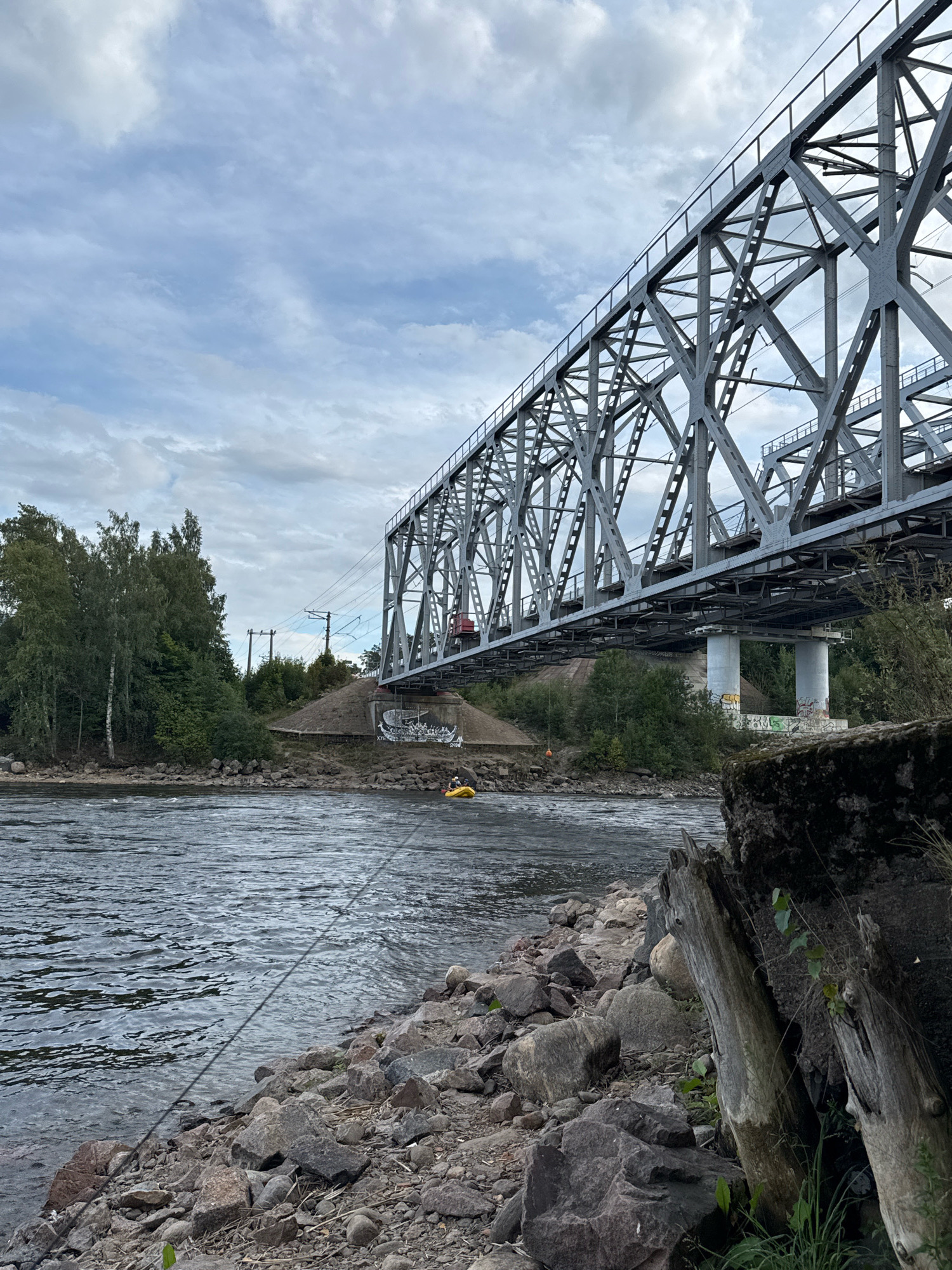 In Losevo, Russia, a wide, dark river flows towards the viewer, its surface rippled by wind. The riverbank is strewn with large, grey rocks and some sparse vegetation. In the foreground, a thin, dark line, possibly a rope or cable, stretches diagonally across the water from the bank. On the opposite bank, a dense line of green trees forms a natural backdrop. Looming above the trees is a massive, grey steel railway bridge, its intricate lattice structure dominating the upper right portion of the image. Beneath the bridge, supporting pillars are visible, one of which bears graffiti. In the middle distance, a small, bright yellow inflatable raft with two people in it is visible on the river. The sky above is cloudy with patches of lighter grey and white.