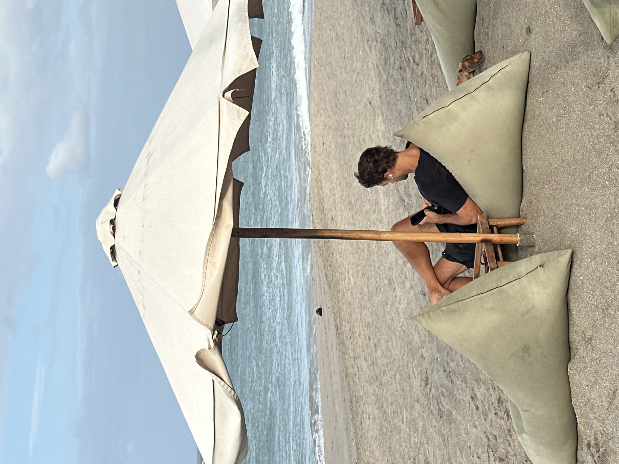 A man is seated under a large, light-colored beach umbrella on a sandy beach in North Kuta, Indonesia. He is wearing a black t-shirt and black shorts, and has white earbuds in his ears. He is holding a black smartphone in his right hand. He is sitting on a small wooden chair which is positioned between two large, triangular, light-green beanbag chairs. The ocean is visible in the background, with waves breaking on the shore. The sky is partly cloudy. A small, brown dog is partially visible between the beanbag chairs on the right side of the image.