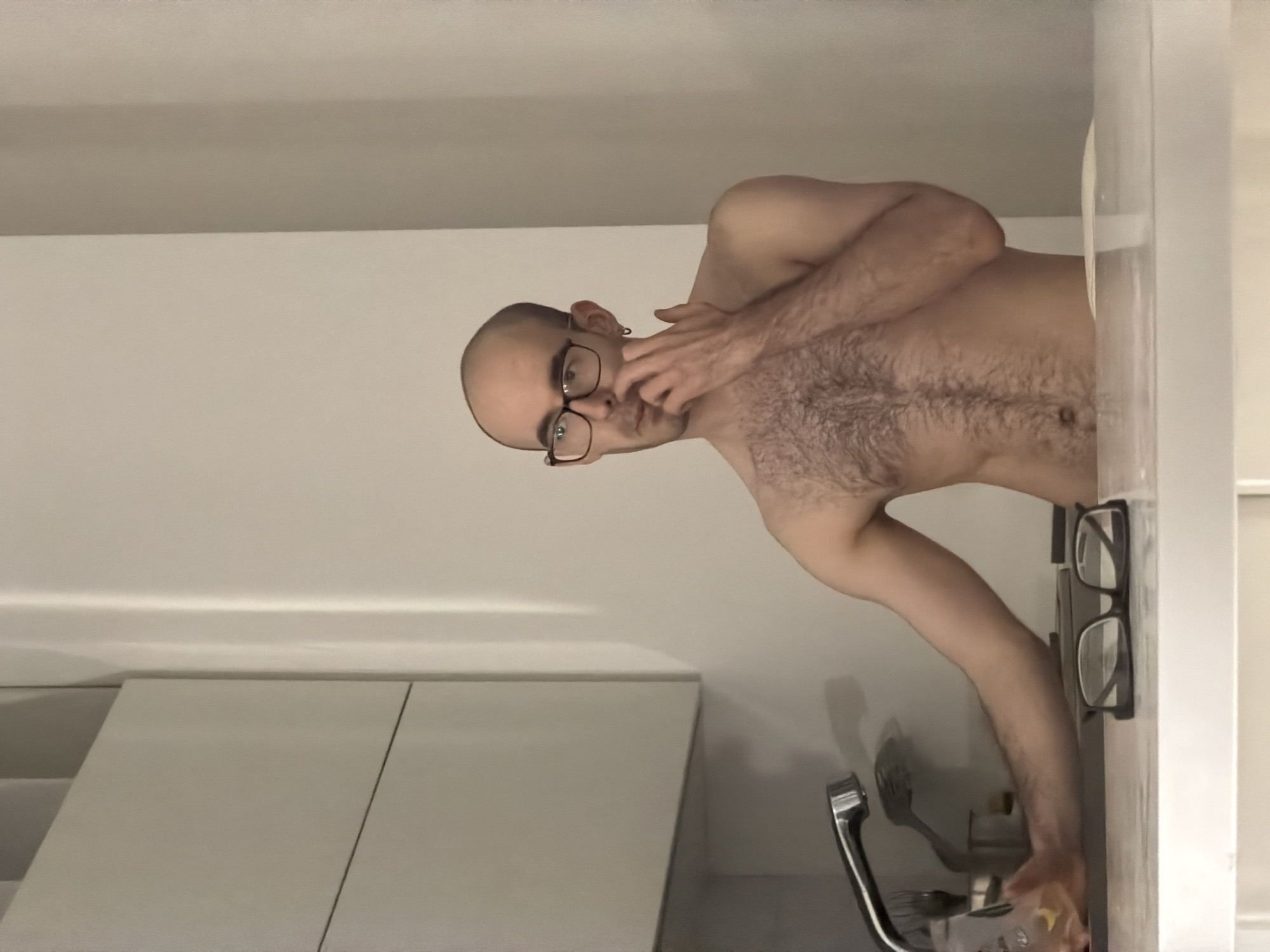 A bald, shirtless person wearing glasses stands in a bathroom or kitchen setting. The person is turned slightly towards the viewer and is touching their face with one hand. Behind the person, there are light-colored cabinets and a chrome faucet. Another pair of glasses rests on a nearby vertical surface. The location is not discernible from the image and therefore cannot be identified as Khimki, Russia.