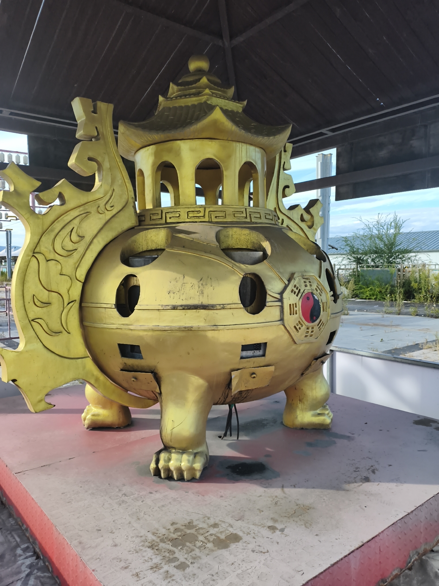 A golden, ornate, incense burner shaped like a mythical creature or a traditional Chinese temple sits on a red-trimmed platform in an outdoor setting. The incense burner is elaborately decorated with swirling motifs on its sides and has multiple openings, resembling windows. A prominent symbol on its side features a red and black yin-yang emblem within an octagonal frame, surrounded by trigrams. The creature's body is supported by four clawed feet. Above the incense burner, a dark wooden roof structure is visible, suggesting it is housed in an open pavilion. The background shows an open, somewhat overgrown area with a building and trees under a light blue sky. The platform and surrounding ground appear to be made of concrete with some signs of wear and dirt. No people are present in the image. The scene is located in 新城村 (Xincheng Village), China.