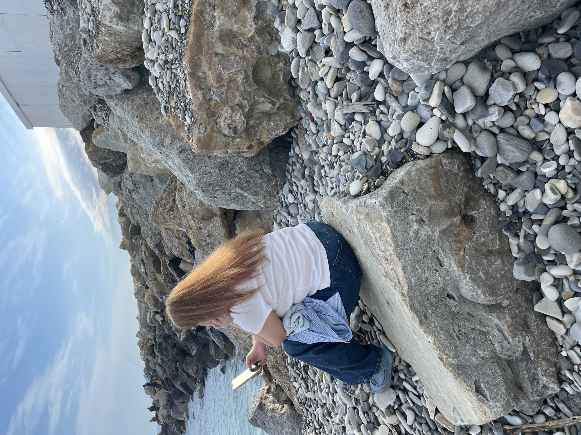 A woman with long, reddish-blonde hair, wearing a white t-shirt and blue jeans, is sitting on a large gray rock at what appears to be a beach or shoreline in Olginka, Russia. She is holding a mobile phone in her right hand and looking down at it. She has a blue shirt bunched on her lap. The ground is covered with smooth, rounded gray and white stones of varying sizes. The background shows other large rocks and the ocean and sky. People are visible on the shore in the background. The scene is captured outdoors under daylight conditions.