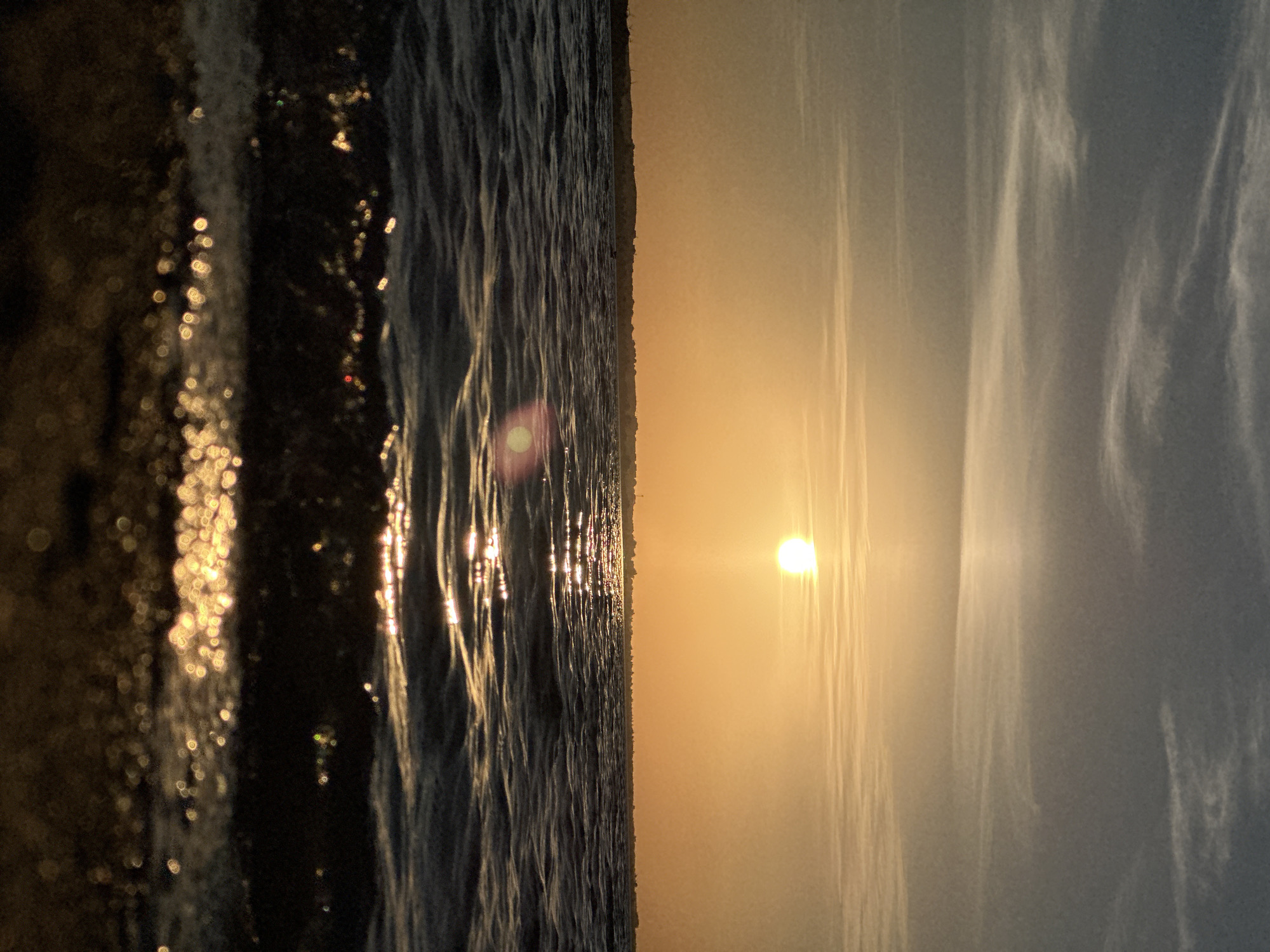 The image depicts a sunset over a body of water, possibly a lake or sea, with a distant shoreline visible. The sun is a bright, luminous orb positioned in the upper center of the frame, casting a warm, golden light across the scene. Wispy clouds stretch across the sky, tinged with hues of orange and yellow from the setting sun. The water in the foreground is rippled, with sunlight reflecting off its surface, creating a shimmering effect. The very bottom of the image shows a dark, textured foreground, likely the shore or edge of the water. There are no people or specific man-made objects clearly discernible in the image, with the exception of a faint, dark line representing the distant shoreline, possibly dotted with structures. The scene is tranquil and natural, evoking a sense of peace and beauty associated with a sunset. The location is identified as the city of Kokshetau, Kazakhstan.