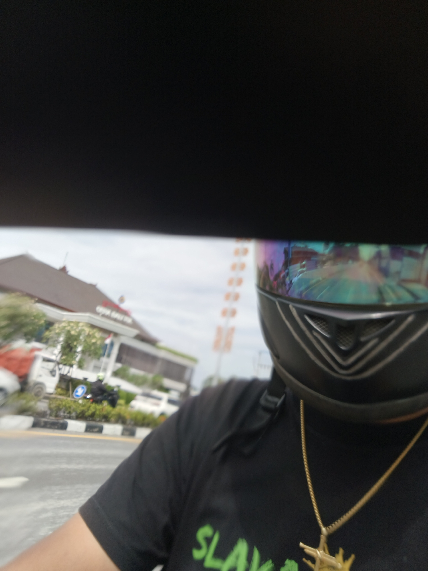 The image shows a person wearing a black t-shirt, a gold chain with a pendant, and a black motorcycle helmet with a reflective visor. The background is blurred, but appears to be an outdoor scene with buildings and vehicles. Based on the context of Denpasar, Indonesia, the image could be taken on a street or near a building in the city. The reflective visor of the helmet shows a distorted image of the surroundings, which might include a road, other vehicles, and buildings. The person is likely riding a motorcycle or is a passenger on one. The black t-shirt has some green lettering on it, but it is difficult to read due to the blur. The overall scene suggests a casual outdoor activity in an urban environment.