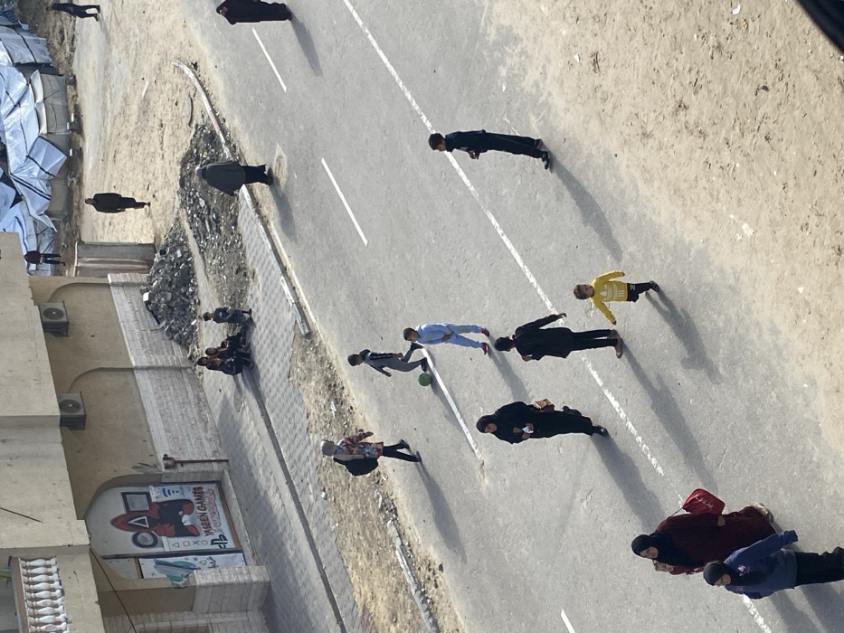 An overhead view captures a street scene in Khan Yunis, Palestinian Territory, under bright daylight, evidenced by distinct long shadows. The primary subject is an asphalt road, marked with two parallel white dashed lines, occupied by several pedestrians.
On the road, two boys are interacting with a green soccer ball; one boy in a light blue tracksuit kicks the ball, while another in a grey top observes. Further left, a woman in a black abaya and head covering walks, carrying a small object. To her right, a young child in a yellow Adidas-branded long-sleeve shirt walks right. A youth in a black top and dark pants stands nearby. In the lower left, a woman in a burgundy abaya holding a red bag walks alongside a girl in a blue jacket and dark head covering. Another woman in dark clothing walks further up the road, heading right.
To the left of the road, a paved sidewalk abuts a multi-story, light brown building with a white-balustered balcony on an upper floor. The ground floor features a storefront with a large "YASEEN GAMES" sign, displaying a red "Squid Game" figure, PlayStation logo, and other gaming icons. Two outdoor air conditioning units are mounted on the building wall above the store. A group of approximately five children are seated on the sidewalk near the storefront.
To the right of the road, the ground transitions to an unpaved, sandy, and dirt-covered area. Piles of debris and rubble are scattered across this unpaved section. In the background, several temporary structures constructed from white tarpaulins are visible, suggesting a camp or temporary settlement. Multiple individuals are seen walking across the dirt expanse and near these temporary shelters.