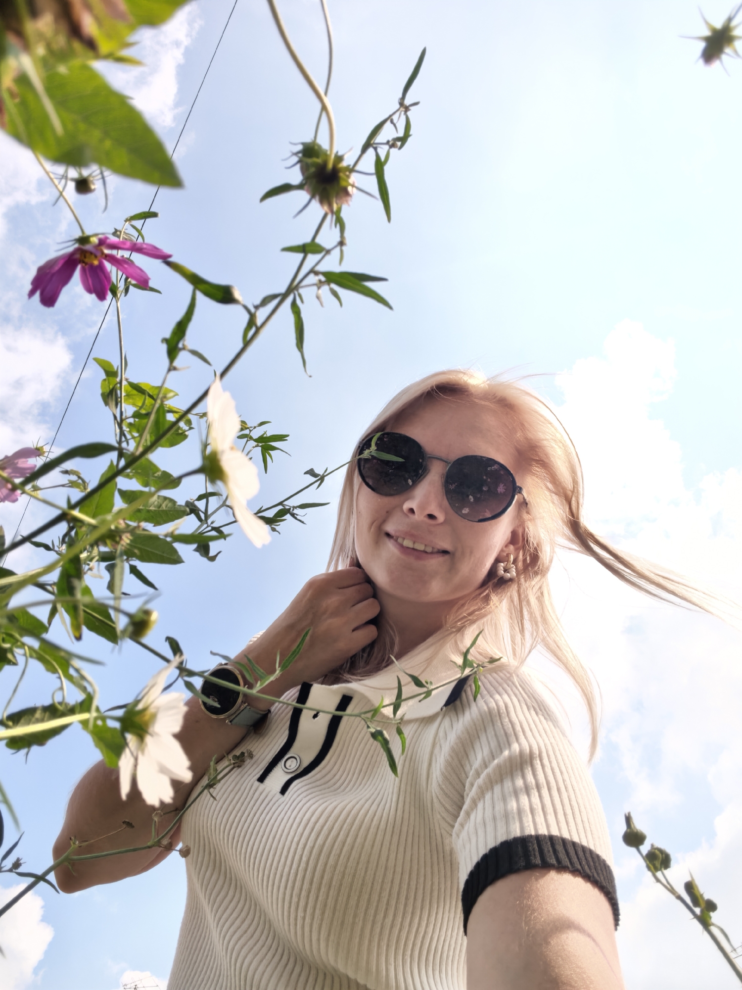 A woman is captured in a low-angle, outdoor, medium shot, looking upwards and smiling at the camera. She has blonde hair, wears round, dark sunglasses, and a white ribbed sweater with black trim on the collar and sleeves. Her left hand is near her neck. A wristwatch with a light blue band is visible on her left wrist. The background consists of a bright blue sky with scattered white clouds and green plants with pink and white flowers, suggesting a garden or park setting. There is no indication of the city of Sterlitamak, Russia.