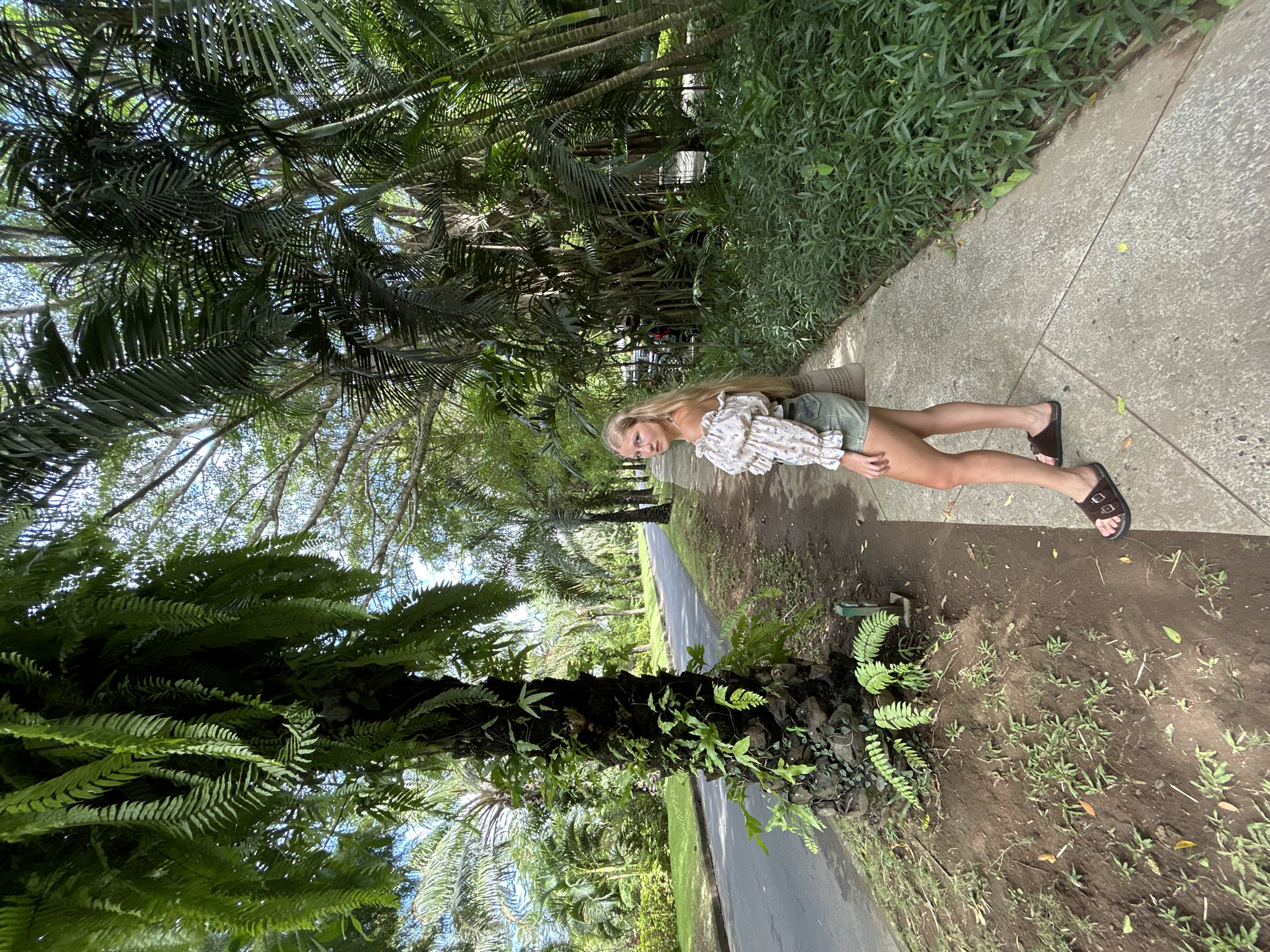 A woman is standing on a concrete path alongside a road in Kuta Selatan, Indonesia. She has long blonde hair and is wearing a floral top and shorts. Her feet are clad in brown sandals. The path is bordered by lush green vegetation and palm trees. A large tree trunk covered in ferns is on the left side of the frame. The road is visible in the background, lined with more trees. The sky is visible through the trees.
