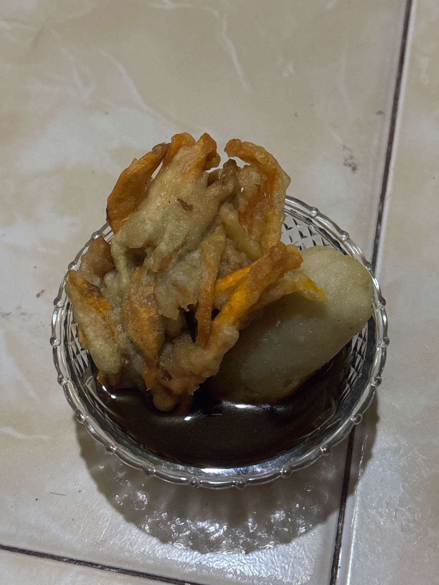 A close-up, overhead shot shows a clear glass bowl filled with a dark liquid and a portion of food. The food consists of several pieces of fried, golden-brown battered items that resemble flower petals, with some visible orange interior. A light-colored, oval-shaped object, possibly a dumpling or cake, is partially submerged in the dark liquid alongside the fried items. The bowl rests on a tiled surface with a light beige and white marble pattern. The lighting casts a soft shadow to the right of the bowl. No people or specific city features are visible.