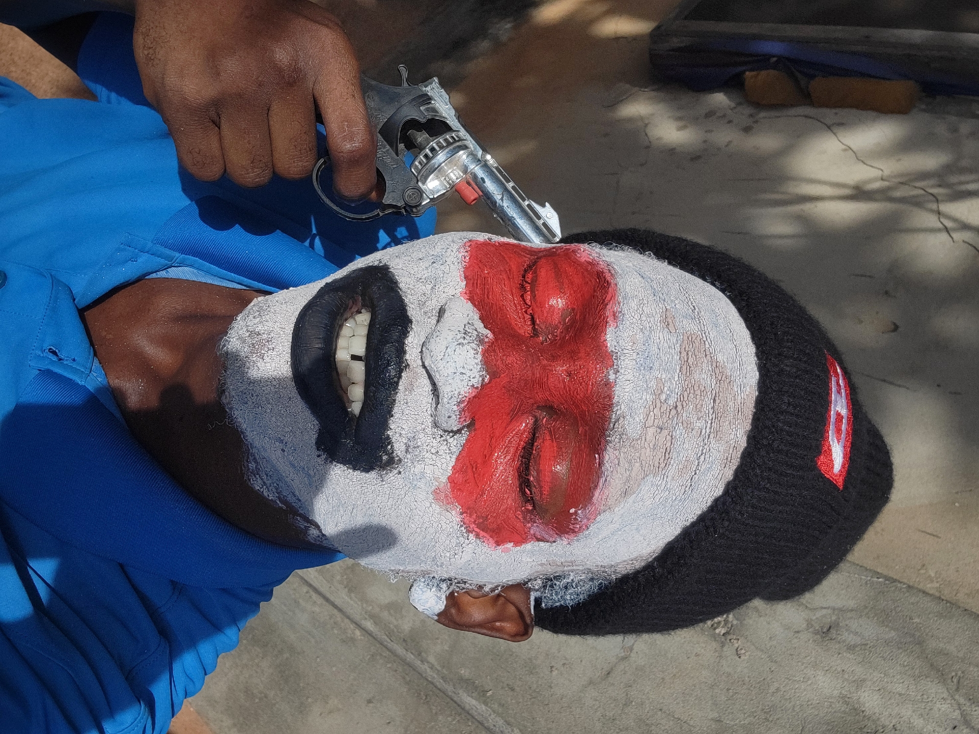 A person with dark skin is lying on their back, wearing a black knit cap with a red and white logo, and has white, red, and black face paint. A hand is holding a silver toy revolver near the person's head. The scene is located outdoors, possibly in Nkpor, Nigeria, as specified. The ground is concrete.