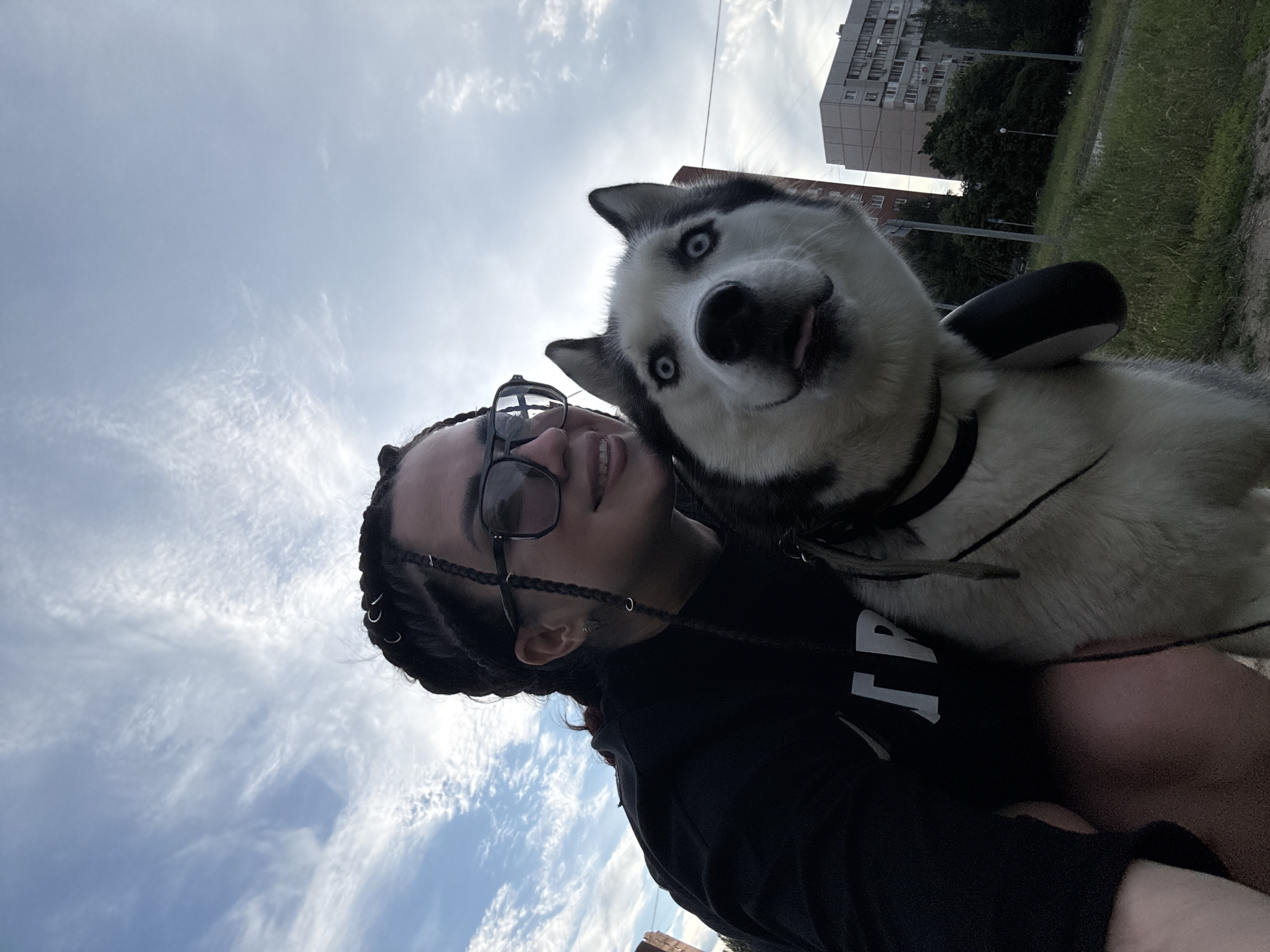 An individual with braided hair and sunglasses is taking a selfie outdoors with a husky. The individual is wearing a black hoodie with white lettering. The husky, positioned in the foreground, has blue eyes and its tongue is slightly out. The background shows a cloudy sky and apartment buildings. The scene is outdoors with grass and trees.