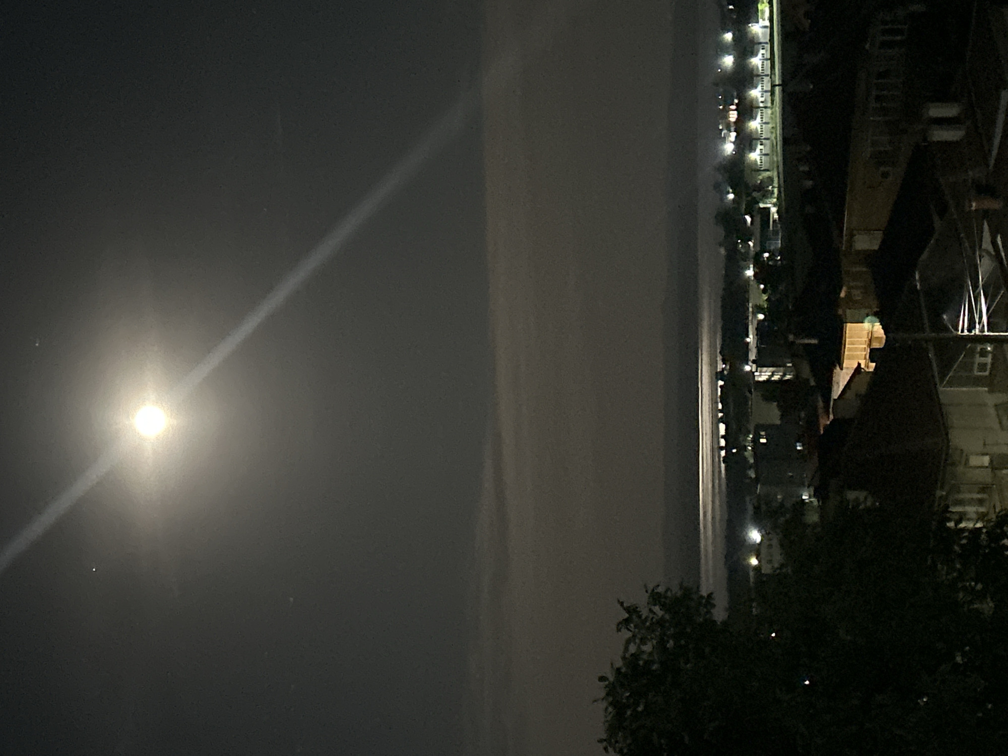 A nighttime view of Burgas, Bulgaria features a bright moon dominating the left side of the frame, its light refracting in a streak. The silhouette of buildings and foliage lines the right side of the image. Lights from other buildings and street lamps are visible in the distance. A dark body of water, possibly a river or the sea, separates the moonlit sky from the urban landscape.