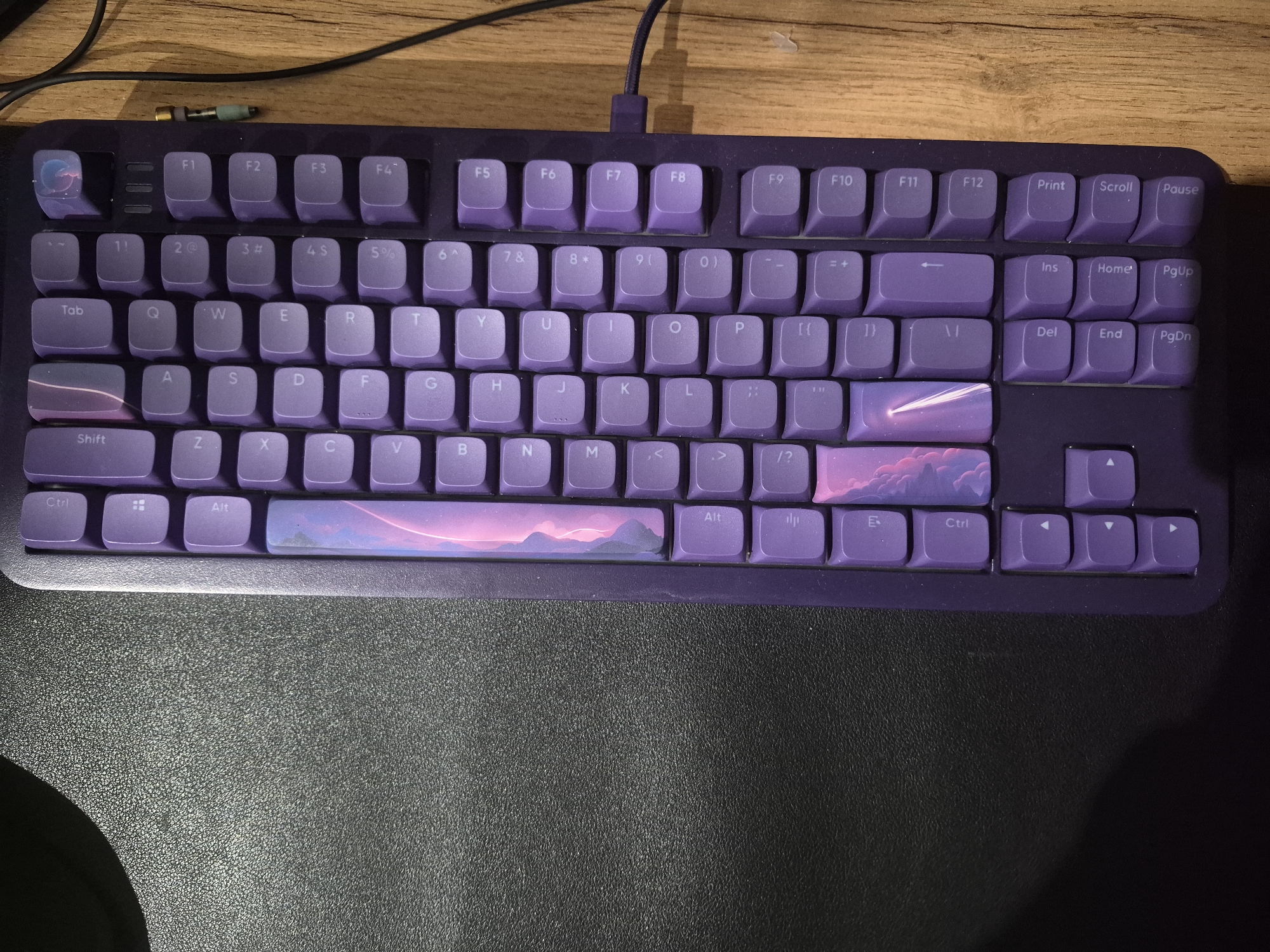 The image shows a close-up, overhead view of a purple mechanical keyboard on a wooden desk. The keyboard is the main subject, with its keys illuminated by a soft light source, casting subtle shadows. The keys are a uniform purple color, with the exception of the spacebar and some of the function keys which feature a custom design. The spacebar and several other keys display a vibrant, stylized image of a sunset or sunrise over a mountainous landscape with a shooting star. The keys are neatly arranged in a standard QWERTY layout, with function keys (F1-F12) along the top, a number row, and the main alphanumeric keys below. The keyboard is connected by a black cable that runs diagonally across the top of the image, disappearing off-frame to the left. The background is a wooden surface, suggesting a desk or table, with a black, textured surface visible in the foreground, likely a mousepad or desk mat. There are no people visible in the image. The scene is static and focuses solely on the keyboard as an object. The visible text on the keys includes standard letter and number designations, as well as function keys like "Tab," "Shift," "Ctrl," "Alt," "Print," "Scroll," "Pause," "Ins," "Home," "PgUp," "Del," "End," and "PgDn." The overall impression is one of a digital workspace, possibly used for gaming or typing, with a personalized aesthetic due to the custom keycaps.