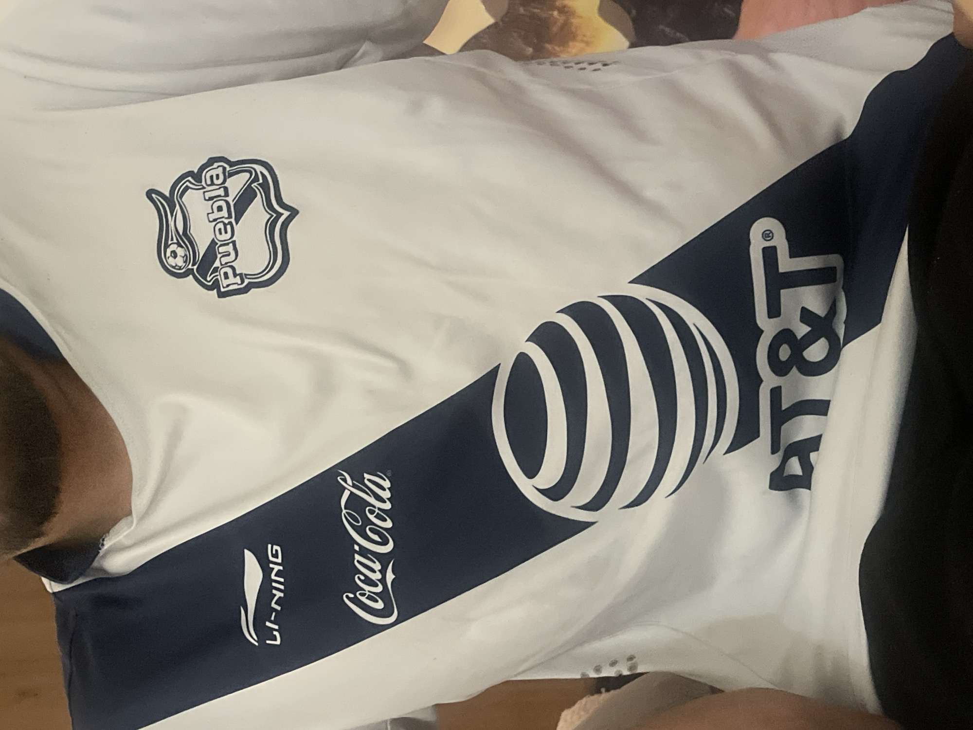 This media file shows a close-up view of a white soccer jersey with navy blue accents. The jersey has several logos and text visible. On the left side of the chest, there is a shield-shaped emblem with the word "Puebla" written inside. A soccer ball is also incorporated into the emblem. Across the chest, a diagonal navy blue stripe features a large, stylized AT&T logo, also in navy blue. Below the AT&T logo, within the blue stripe, is the Coca-Cola logo in white script. On the lower left side of the jersey, also within the blue stripe, is the LI-NING logo and text. The jersey appears to be worn by a person, with part of their shoulder and arm visible on the left side of the frame, and their other arm and side visible on the right, wearing dark clothing. The lighting suggests an indoor setting, possibly a room, as there are no clear outdoor environmental cues. The image is taken from a slightly angled perspective, looking down at the jersey. No specific time of day, weather, or emotional cues are discernible from the image.