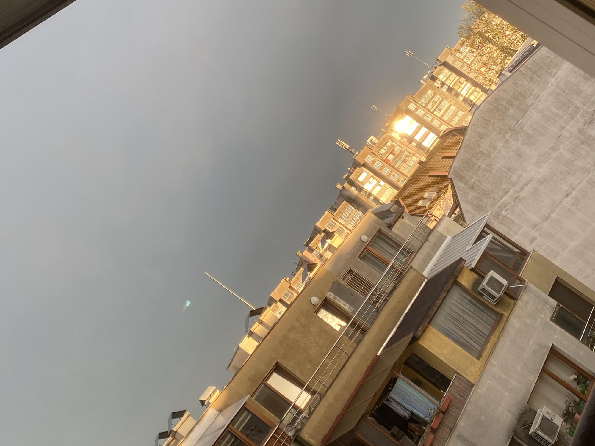 The image, taken from a low angle, depicts a view of several buildings against a grayish sky. The buildings are a mix of older, weathered structures with windows and balconies. The strong sunlight reflects off the windows of the taller building, creating a bright glare. Air conditioning units are visible in some windows. The buildings appear to be located in Varna, Bulgaria.