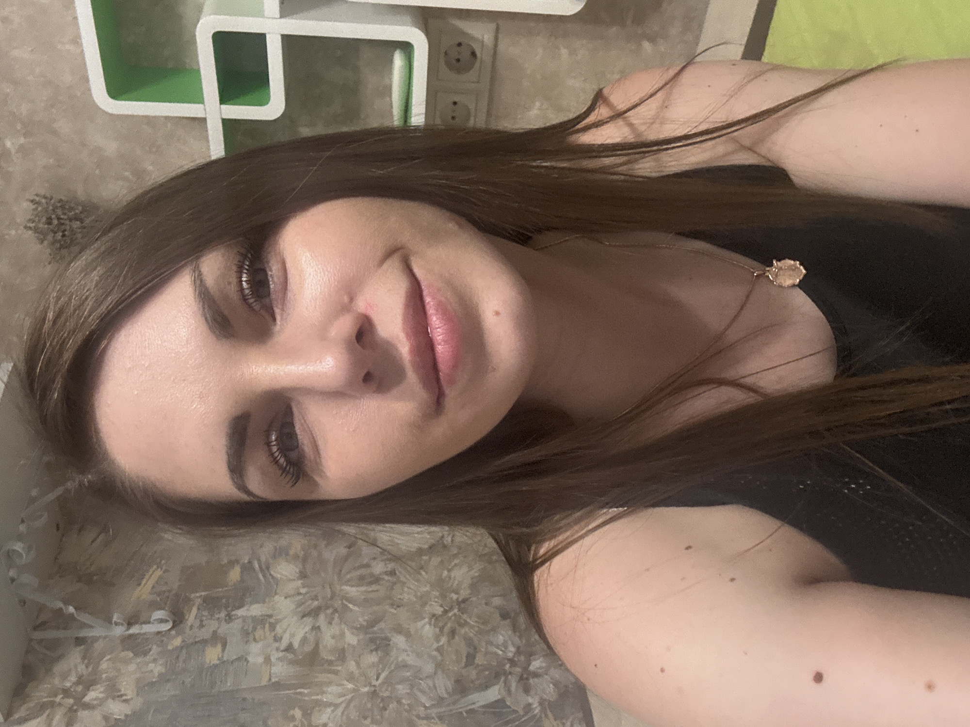A close-up, rotated image depicts a woman with long, brown hair. She is wearing a black top and a delicate gold necklace with a pendant. The background includes a shelf with green and white elements and an electrical outlet. The location information is not discernible from the image.