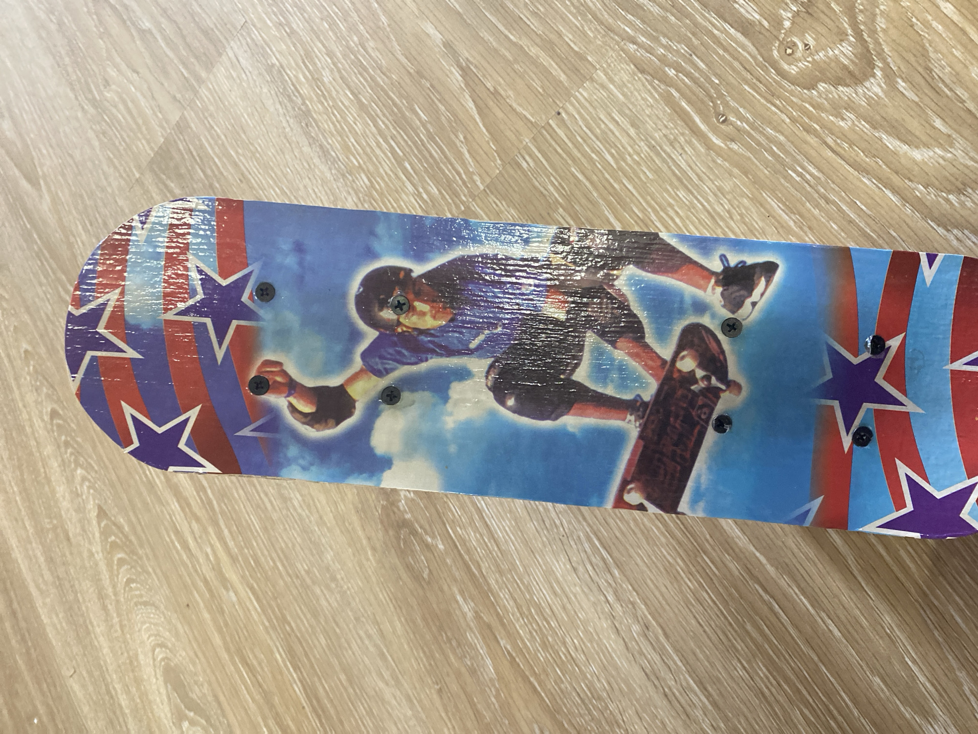 The media file displays the top surface of a skateboard resting on a light-colored wooden floor in Saint Petersburg, Russia.
The primary subject is a skateboard, featuring a vibrant graphic design. The central part of the skateboard depicts a male skateboarder in mid-air, performing a trick against a blue sky with white clouds. The skateboarder is wearing a blue short-sleeved shirt, dark shorts, a helmet, and what appear to be knee pads and wrist guards. His arms are extended, and he is holding the skateboard with one hand. The ends of the skateboard are decorated with a patriotic-themed pattern, consisting of alternating red and blue stripes, bordered by white outlines, and interspersed with purple stars.
Several black Phillips head screws are visible on the surface of the skateboard, indicating the mounting points for the trucks. The graphic surface itself appears to have a protective layer that reflects light, creating horizontal streaks across the image of the skateboarder. The skateboard's edges show some minor wear.
The skateboard is laid horizontally on a light brown wooden or laminate floor, characterized by distinct wood grain patterns running mostly perpendicular to the length of the board. The lighting is bright and even, suggesting an indoor setting. No activity is taking place within the captured scene, as the skateboard is stationary. There is no visible text on the skateboard or the floor.