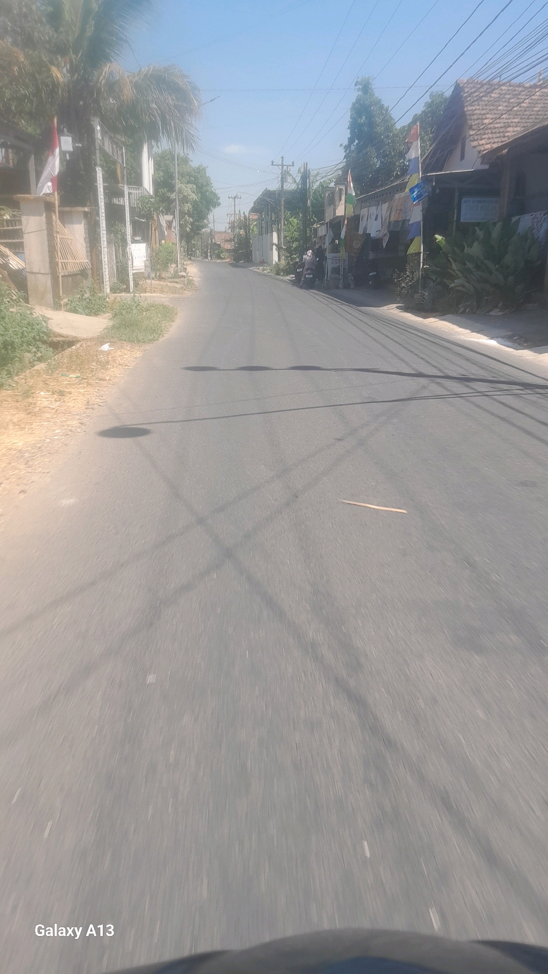The image depicts a street scene in Tenggeles, Indonesia, under a clear blue sky. The road, a paved asphalt surface, is the central focus, stretching into the distance. On either side of the road are residential buildings and vegetation. On the left, there's a palm tree and what appears to be a fence or wall structure with a small Indonesian flag. Further down the street, on the right, houses with traditional tiled roofs are visible. Several utility poles and overhead wires crisscross the scene. Shadows from the poles and wires are cast across the road, creating a pattern of light and dark. Two motorcycles are parked on the right side of the road near one of the houses. The overall impression is of a quiet, sun-drenched street in a rural or semi-urban Indonesian setting. The image also includes the text "Galaxy A13" in the bottom left corner, indicating the device used to capture the photo.