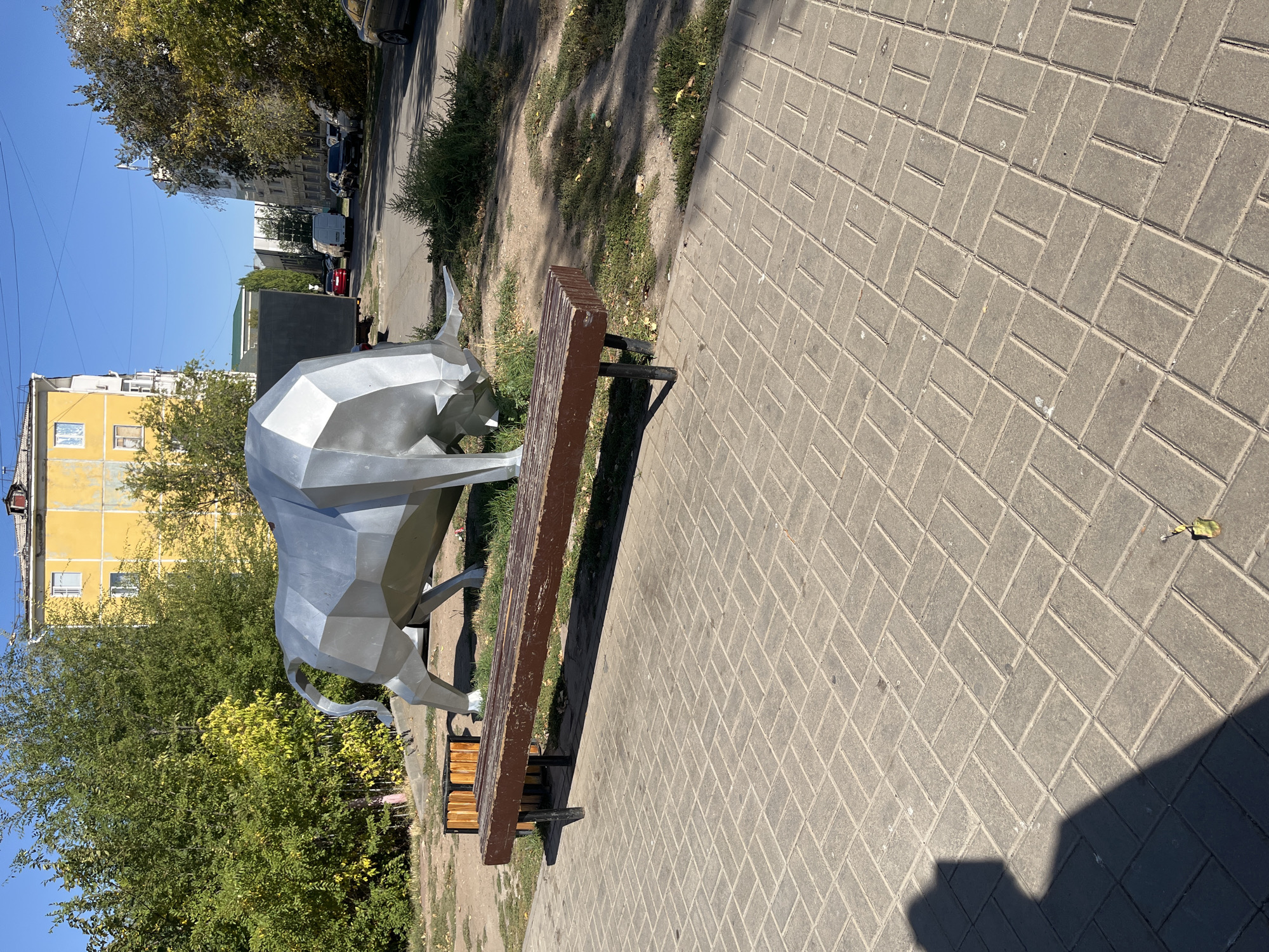 A low-polygon silver sculpture of a bull stands on a paved area, facing left. The sculpture is in a park-like setting, with trees and grass surrounding it. Behind the sculpture, a wooden bench is situated on the paved area. In the background, there is a yellow apartment building, other buildings, and a street with cars. The sky is clear and blue. The scene appears to be in Astana, Kazakhstan.
