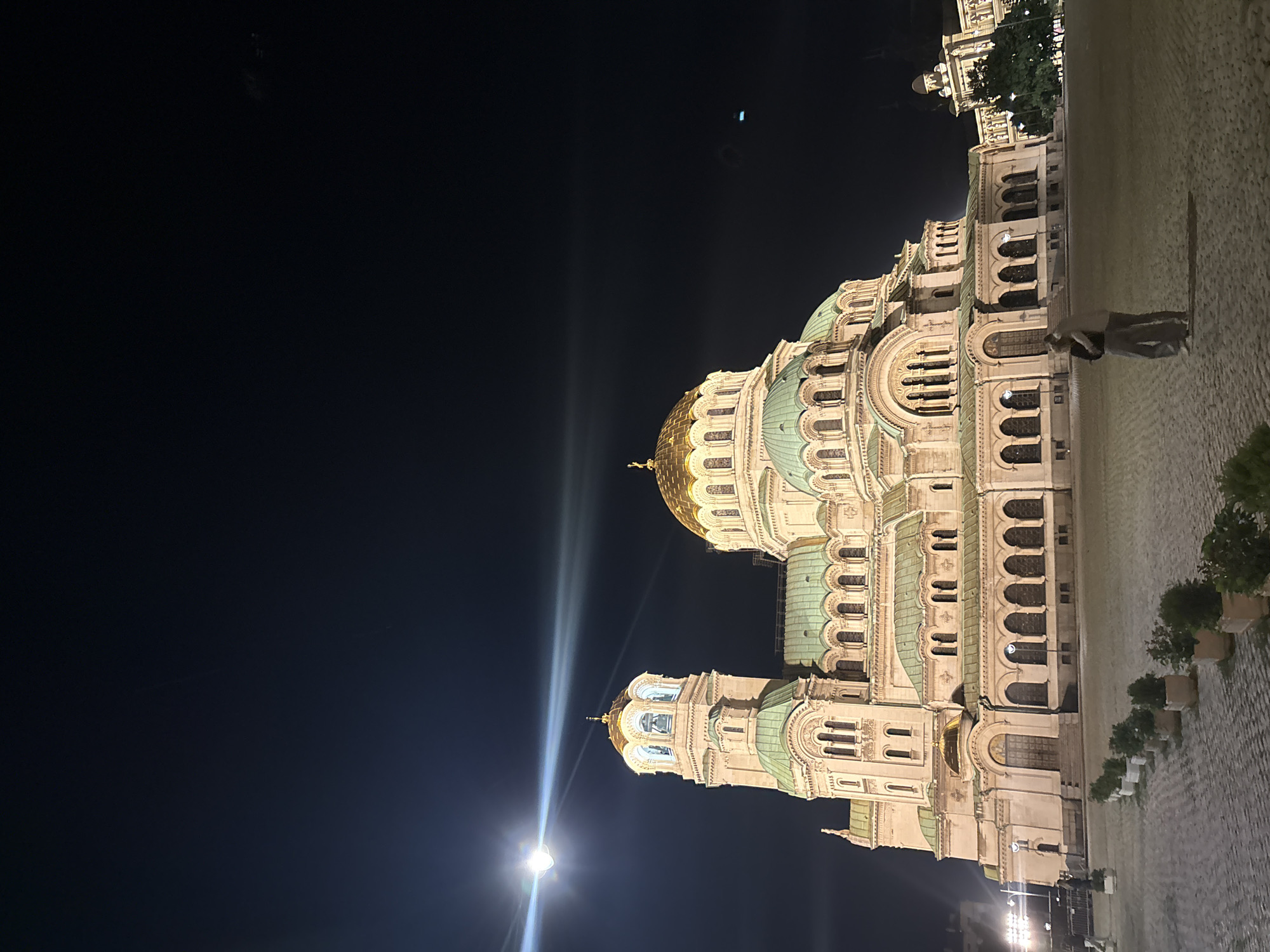 The image displays the Alexander Nevsky Cathedral in Sofia, Bulgaria, at night. The grand Orthodox cathedral dominates the right side of the frame, illuminated against a very dark, clear sky. Its distinctive architecture features a prominent central golden dome and several smaller, green-domed towers. The facade is constructed from light-colored stone, adorned with intricate details, arched windows, and columns, all highlighted by artificial lighting.
In the foreground, a wide, cobblestone-paved square extends towards the cathedral. A lone person is visible walking on the cobblestones near the building, appearing as a blurred figure. On the far right, a line of potted plants or small shrubs is positioned along the edge of the paved area.
Bright light sources, likely streetlights, create a strong glow and lens flares in the lower-left corner of the image, streaking across the dark sky. Additional circular lens flares or ghosting are visible higher in the dark sky. The scene depicts a serene nighttime view, emphasizing the majestic presence of the cathedral. There is no visible text in the image.