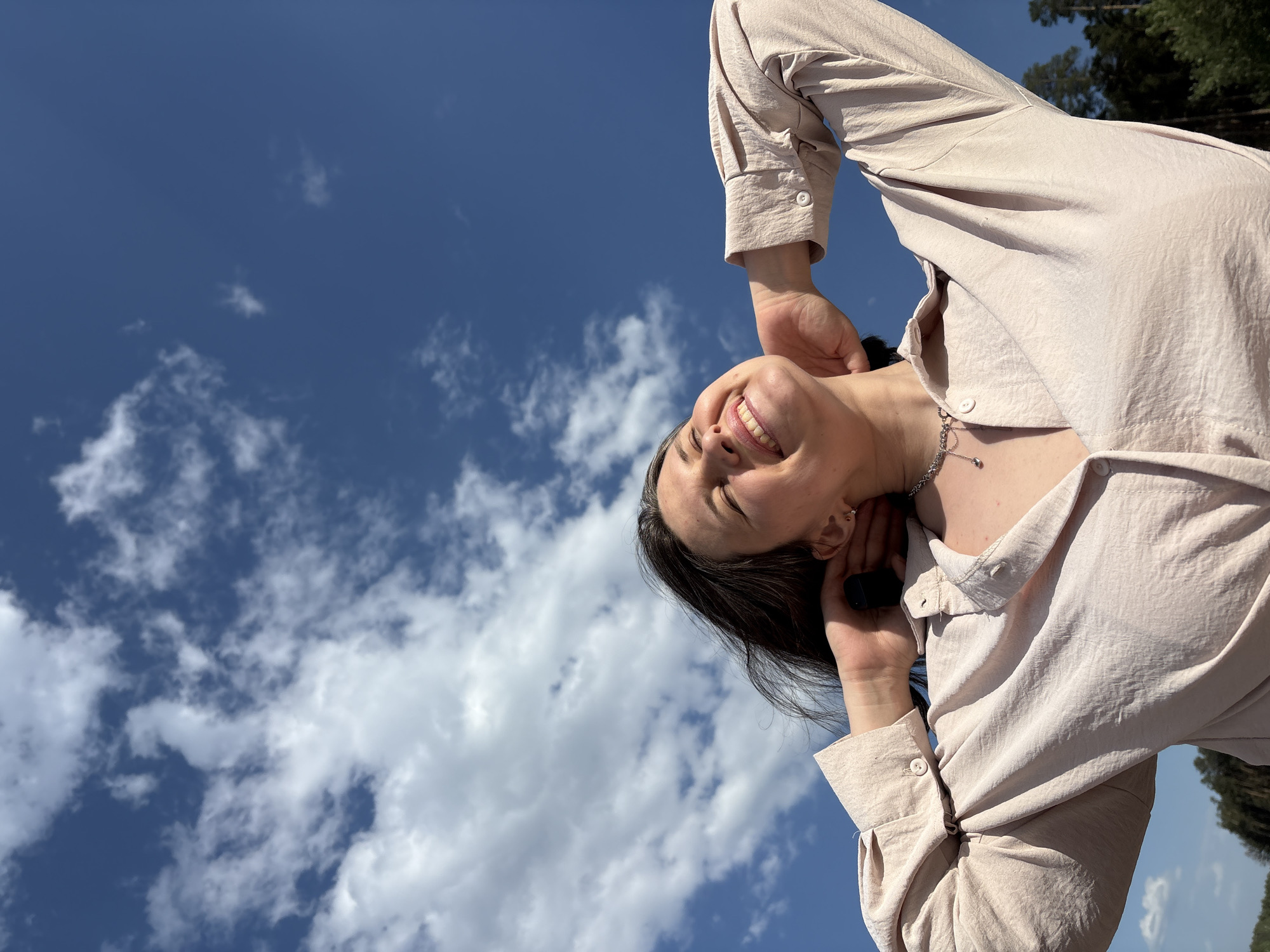 A woman is smiling up at the sky, her hands are behind her head, and she is wearing a light-colored shirt. The sky is blue with scattered white clouds. The location appears to be Chaikovsky, Russia. There are treetops visible in the background. The woman is also wearing a necklace.