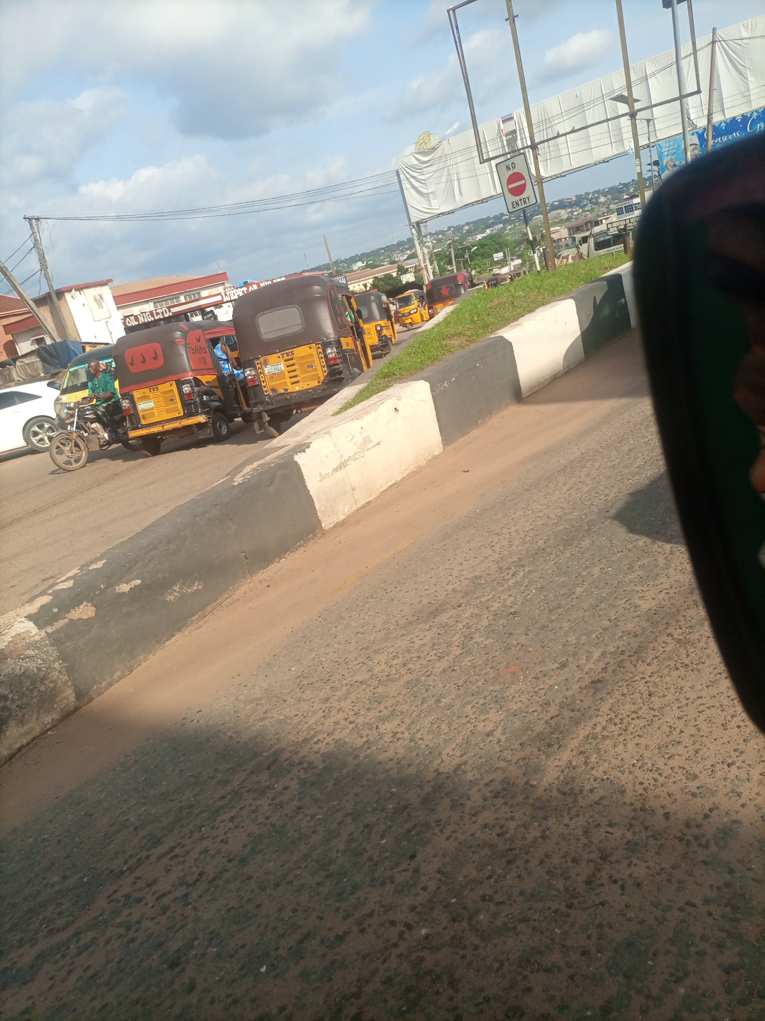The image captures a street scene in Nkpor, Nigeria, featuring a line of auto-rickshaws, known locally as Keke Napep, parked or moving along the road. The vehicles are primarily yellow, with some exhibiting brown canopies. One Keke Napep has the number "118" visible on its side. A man is seen riding a motorcycle in the foreground, and a white car is parked on the left side of the road. Buildings, including one with the signage "OIL NIG. LTD.", are visible in the background. A "No Entry" sign is posted on a pole above the road. The scene is set against a backdrop of a cloudy sky and a generally urban environment with some greenery. The perspective suggests the photo was taken from within another vehicle.