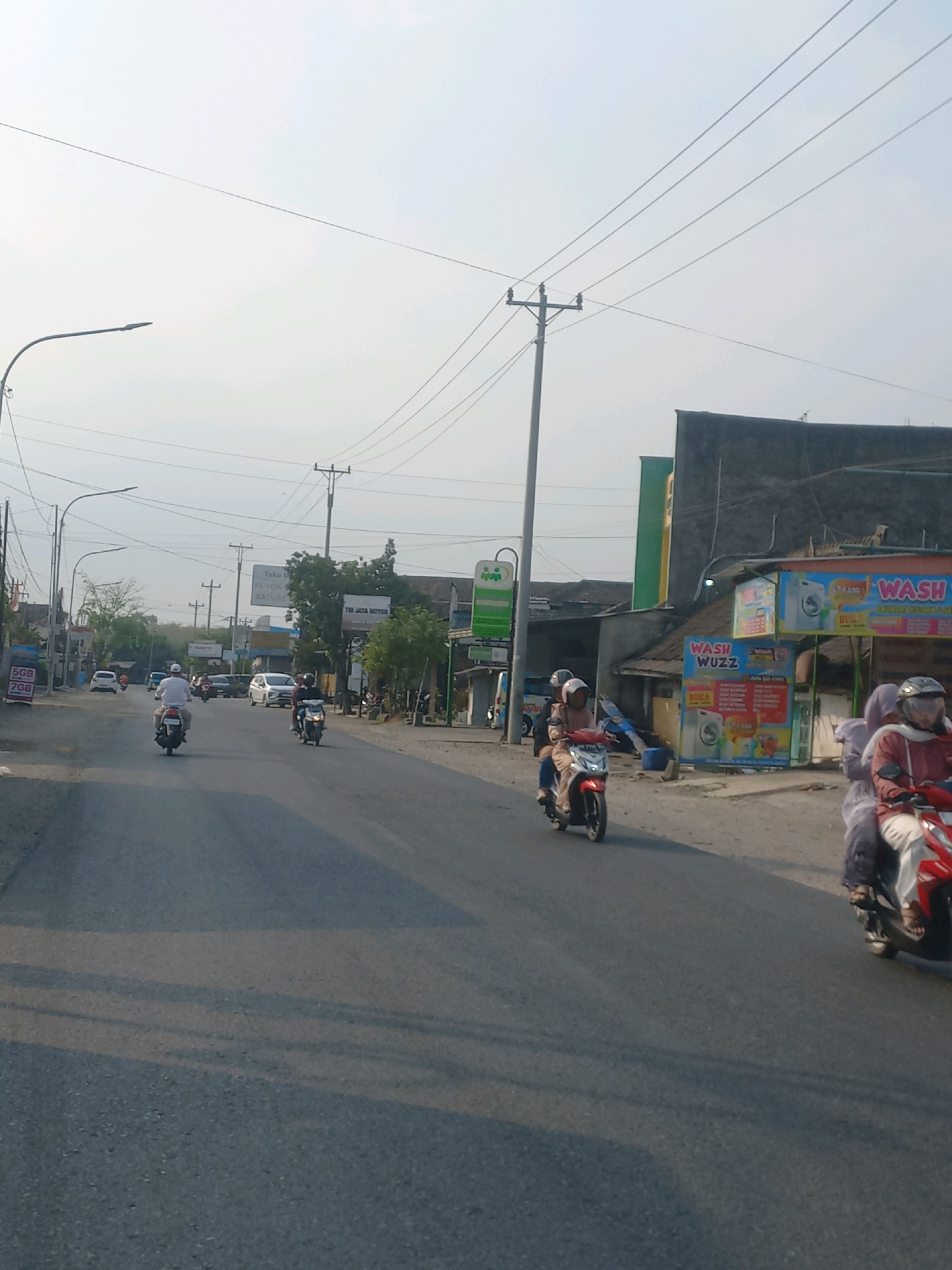 The image depicts a street scene in Baturetno, Indonesia, bathed in the soft light of what appears to be late afternoon or early morning. The sky is overcast and pale, with power lines stretching across it. On the left side of the road, a modern street lamp with a curved arm is visible. The road itself is paved with asphalt and marked with yellow lines.
Several people are seen riding motorcycles. In the foreground, on the right, a person wearing a red jacket and white pants rides a red motorcycle, with a passenger behind them. To the left, a person in a light-colored shirt and white pants rides a dark motorcycle, also with a passenger. Further down the road, more motorcycles are visible, with riders and passengers. A white car is also present in the distance.
Along the roadside, various buildings and structures are present. On the right, a building with a dark facade is topped with a colorful sign for "WASH WUZZA" and "WASH". Another sign, possibly for a business, is also visible. On the left, a sign for "TOKO" and "JIYA JAYA MOTOR" is mounted on a pole, alongside smaller signs advertising phone data plans. Greenery, including trees and bushes, is scattered throughout the scene, particularly behind the roadside structures. Utility poles with wires are a common feature, interspersed with street lights. The overall impression is of a moderately busy street in a residential or commercial area.