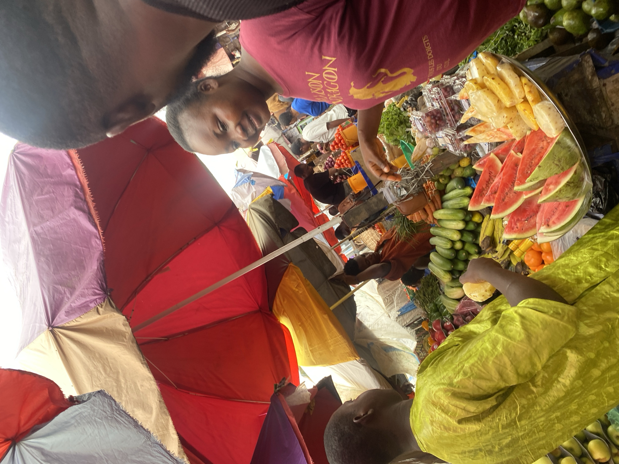 A crowded market scene in Ado, Nigeria, is captured from a low angle under colorful umbrellas. A woman in a maroon shirt with gold lettering and a lion logo smiles at the camera as she arranges produce on a large tray, including watermelon, pineapple, cucumbers, and carrots. Another person in a lime green shirt is selecting a piece of pineapple. Above, portions of two other people's heads are visible: one obscured at the top, and another at the bottom right. Various other merchants and customers are partially visible in the background amongst market stalls and colorful awnings.