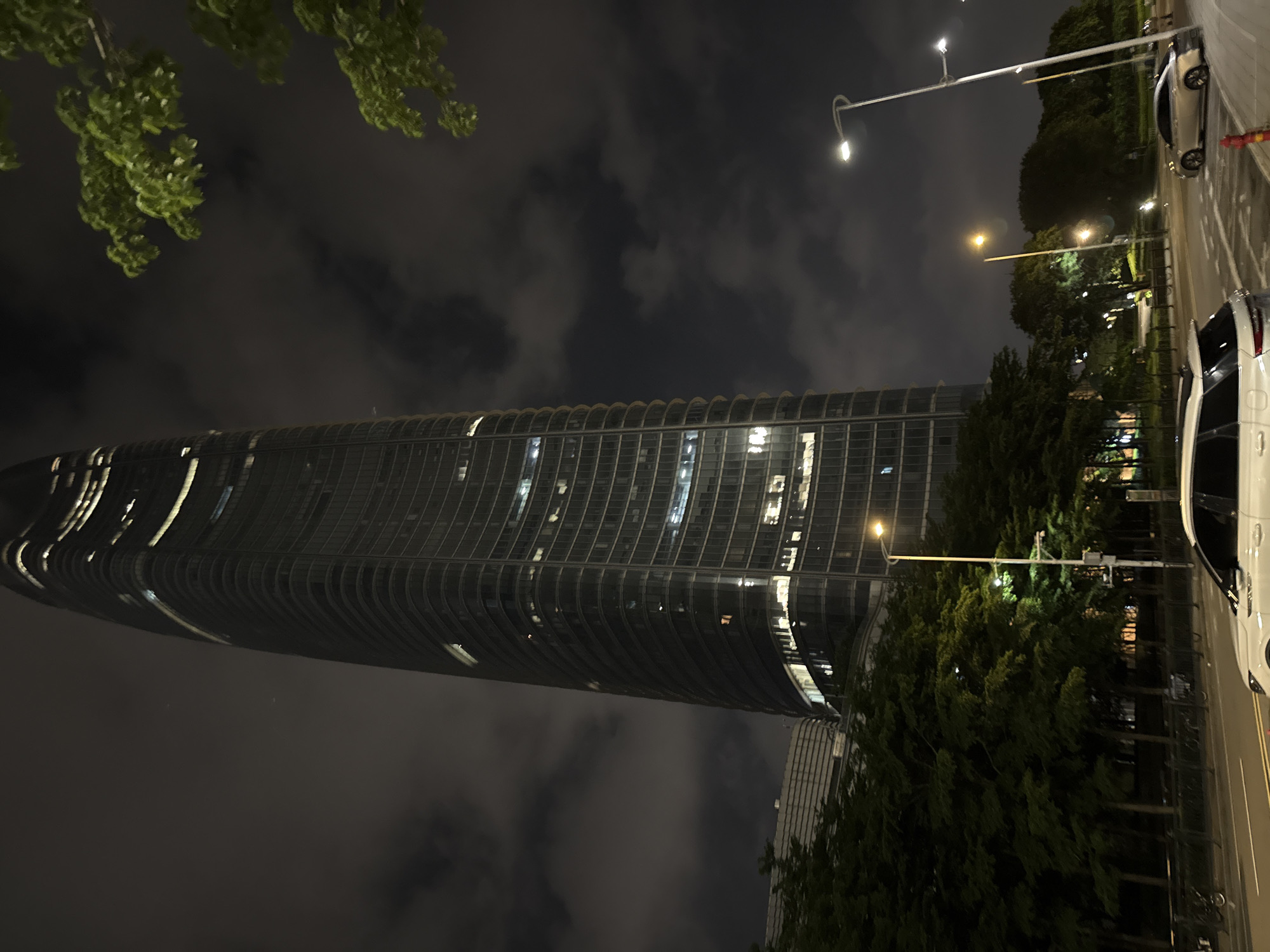 This is a nighttime photograph taken in Haizhu District, China. The scene features a modern, curved skyscraper with numerous lit windows, suggesting it is occupied. In the foreground, lush green trees partially obscure the lower portion of the building and frame the upper left of the image. To the right of the skyscraper, a street runs alongside the building, bordered by more trees and a sidewalk. Several cars are visible parked on the street and sidewalk, including a white SUV with its doors open, hinting at recent activity. Streetlights illuminate the scene, casting a warm glow on the pavement and surrounding greenery, and reflecting off the building's facade. The sky is dark and cloudy, characteristic of nighttime. The overall impression is one of urban dynamism and nighttime activity in a well-developed district.