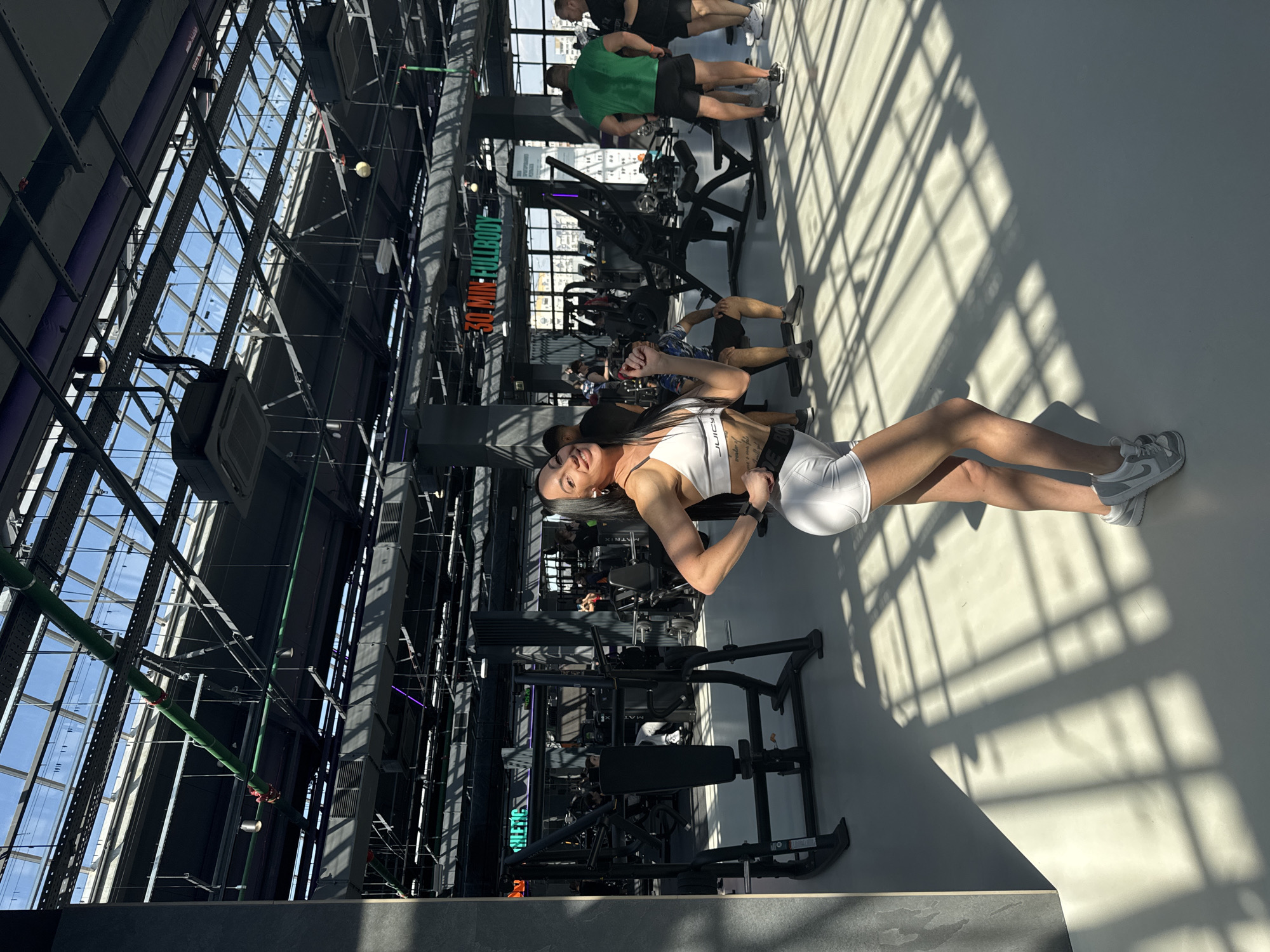 The image captures the interior of a modern fitness facility in Kazan, Russia. The gym features a high ceiling with an elaborate network of metal beams and glass panels that allow natural light to flood the space. Various exercise machines are arranged throughout the area, and several people are engaged in different fitness activities.
A woman in white athletic wear is prominently positioned in the foreground. She is standing and looking towards the camera.