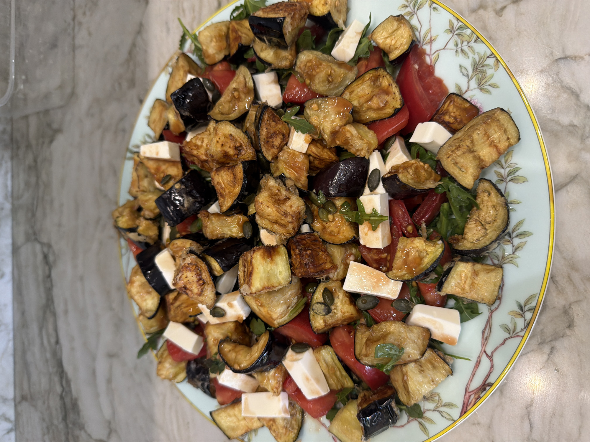 A close-up, eye-level shot captures a generously portioned salad on a decorative pale blue plate with a botanical pattern. The salad is a colorful medley of roasted eggplant pieces, some charred to a deep brown and others a golden-fried hue. Interspersed among the eggplant are bright red cubes of tomato, white cubes of cheese, and scattered green arugula leaves. Several dark green pumpkin seeds are visible throughout the dish, adding a textural element. The plate rests on a speckled marble countertop. The lighting is bright and natural, highlighting the glossy sheen of the roasted vegetables. No people or specific city location are discernible.