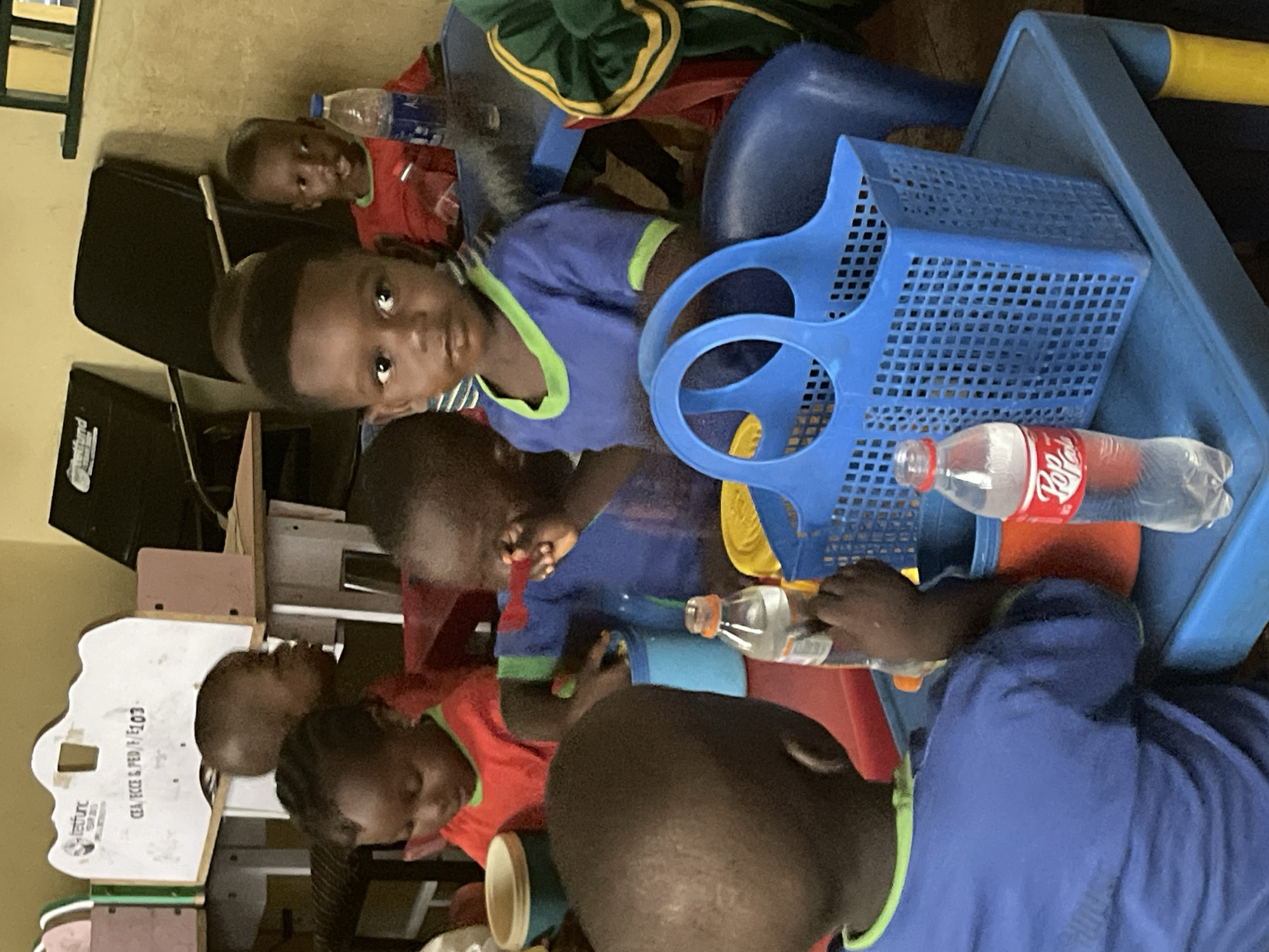 This image captures a group of young children, likely boys, gathered around small blue plastic tables and chairs indoors, suggestive of a classroom or daycare setting in Akwanga, Nigeria. There are at least seven children visible, engaging in an activity.
The subjects include several children with dark skin and short hair. Many wear blue short-sleeved tops with lime green trim around the neck and sleeves, while some wear red tops, also with green trim. One child's attire is green and yellow striped.
Key objects in the scene are the low, blue plastic tables and matching chairs, typical for early childhood education. Several blue plastic baskets, possibly for toys or supplies, are on or near the tables. Multiple clear plastic water bottles are present, one prominently displaying a "Dr Pepper" label with its distinct red branding. One child in a blue shirt is actively holding and looking at a water bottle.
The activity appears to be a focused group session, possibly learning or playtime. Some children are looking directly towards the camera with neutral or slightly curious expressions, while others are engrossed in what's on the table in front of them. The atmosphere seems calm and engaged.
Notable details include the indoor lighting, indicating it is daytime. The children's clothing and the general environment are consistent with the provided location context of Akwanga, Nigeria. On the left side of the image, a white board-like object is partially visible with faint text, including what appears to be "CEAD PICS & PED" and possibly "VENTURE" along with an indistinct logo. Another black piece of furniture is in the background.