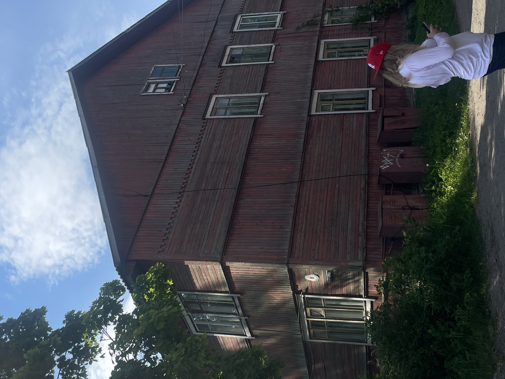 The image depicts an outdoor daytime scene in Priozersk, Russia, identifiable by a street sign on the building. The central subject is a three-story, reddish-brown wooden building with faded paint and weathered vertical planks. It features multiple windows with white frames, some containing sheer curtains, and a dark pitched roof. A circular house number "11" and a rectangular sign inscribed with "Поварская ул." (Povarskaya Street) are visible on the building's left corner, beneath a ground-floor window. At the base of the building, on the right side, are several reddish-brown metal utility boxes, one bearing white graffiti. A black cable runs vertically along the right facade.
To the left, a large tree with abundant green foliage partially obscures the building, with sunlight filtering through its leaves and casting shadows. Lush green bushes and tall grass grow around the base of the building and along the left foreground. A dirt path with scattered grass patches occupies the foreground.
In the lower right foreground, a blonde-haired individual is observed from behind, facing away from the camera. They are wearing a red baseball cap with a white "NY" logo, a light-colored long-sleeved shirt, and dark trousers. The person is holding a smartphone with both hands, appearing to be engaged with the device. The sky is clear blue with scattered white cumulus clouds, and bright sunlight illuminates the scene from the upper left.