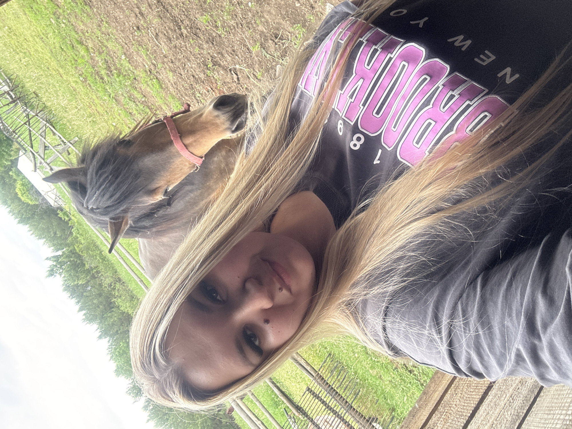 A selfie photograph features a fair-skinned woman with long blonde hair. She wears a dark gray t-shirt with a pink and white "BROOKLYN NEW YORK 1898" graphic. A brown horse wearing a red halter stands to the left of the woman. The background consists of a grassy field enclosed by a wooden fence. The location is unspecified, with no discernible elements indicating it is Большой Букор, Russia.