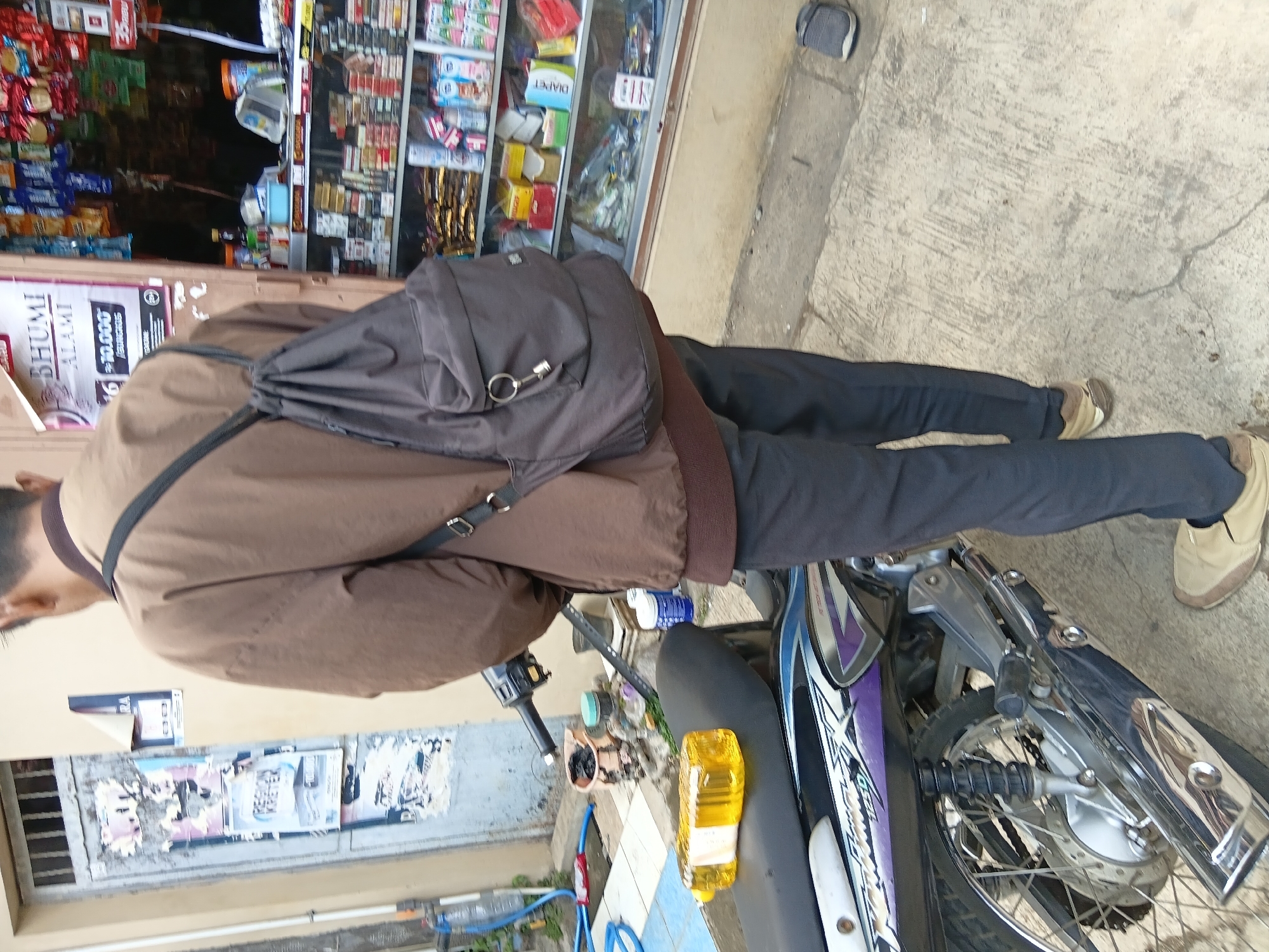 A person wearing a brown jacket, dark pants, and tan shoes stands with their back to the camera in front of a shop entrance in Tanggul Wetan, Indonesia. The person is carrying a dark gray backpack over their left shoulder. To the left of the person, a motorcycle is parked, with a yellow bottle of liquid resting on its seat. The shop behind the person displays various products on shelves visible through a glass window. The ground is paved with concrete.