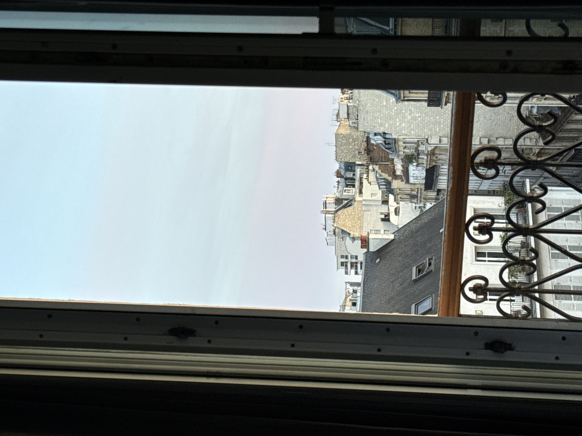 The image is a view from a window looking out over the rooftops of Paris, France. The sky is a pale blue with a hint of pink, suggesting either early morning or late evening. The foreground of the image is dominated by the window frame, with a dark curtain on the left and a grey metallic window frame on the right. A wooden railing with decorative ironwork is visible just below the window. Beyond the railing, rows of Parisian buildings with distinctive mansard roofs, dormer windows, and stone facades stretch into the distance. The architecture is typical of Haussmannian Paris. No people are visible in the image. The overall scene captures a quiet, urban landscape during a calm moment of the day.