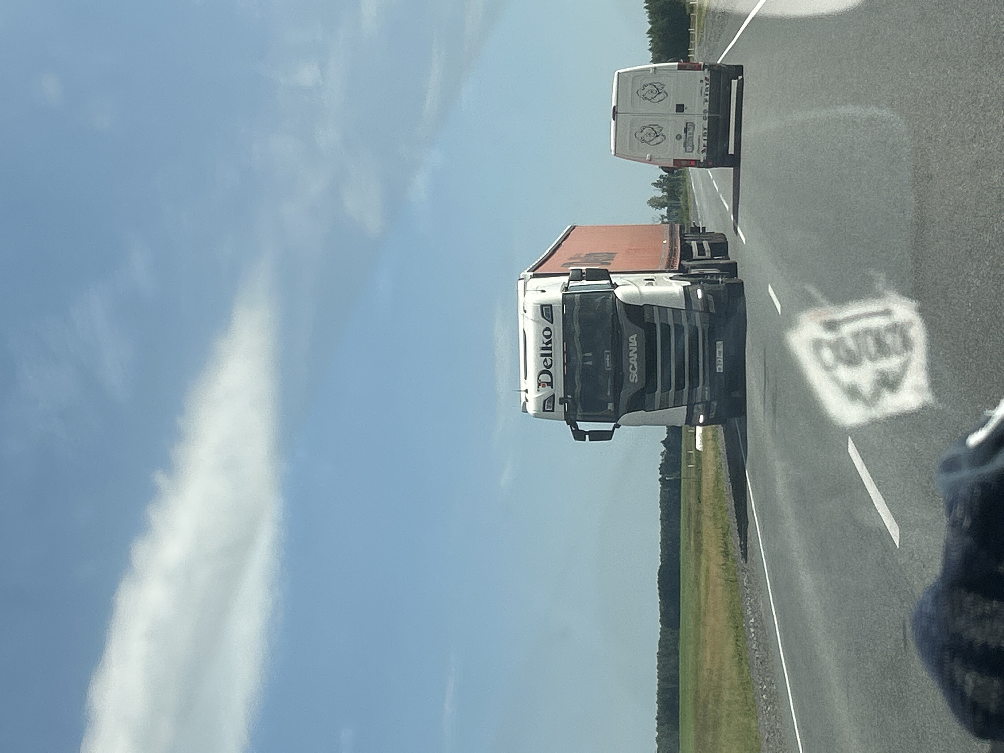 This image depicts a view from a vehicle traveling on a highway, likely in the vicinity of Iliushkinskaya, Russia, given the context provided. The sky is a clear blue with a few wispy white clouds. On the highway, a large white semi-truck with an orange trailer, branded "Delko" and "SCANIA," is in the left lane. In the right lane, a white van with two cartoon bear logos on its rear doors is traveling ahead of the semi-truck. The road surface is asphalt with white lane markings. To the sides of the highway are grassy fields bordered by a line of trees in the distance. The sun is high in the sky, casting shadows. The foreground shows a blurred reflection on the windshield, obscuring parts of the lower image.