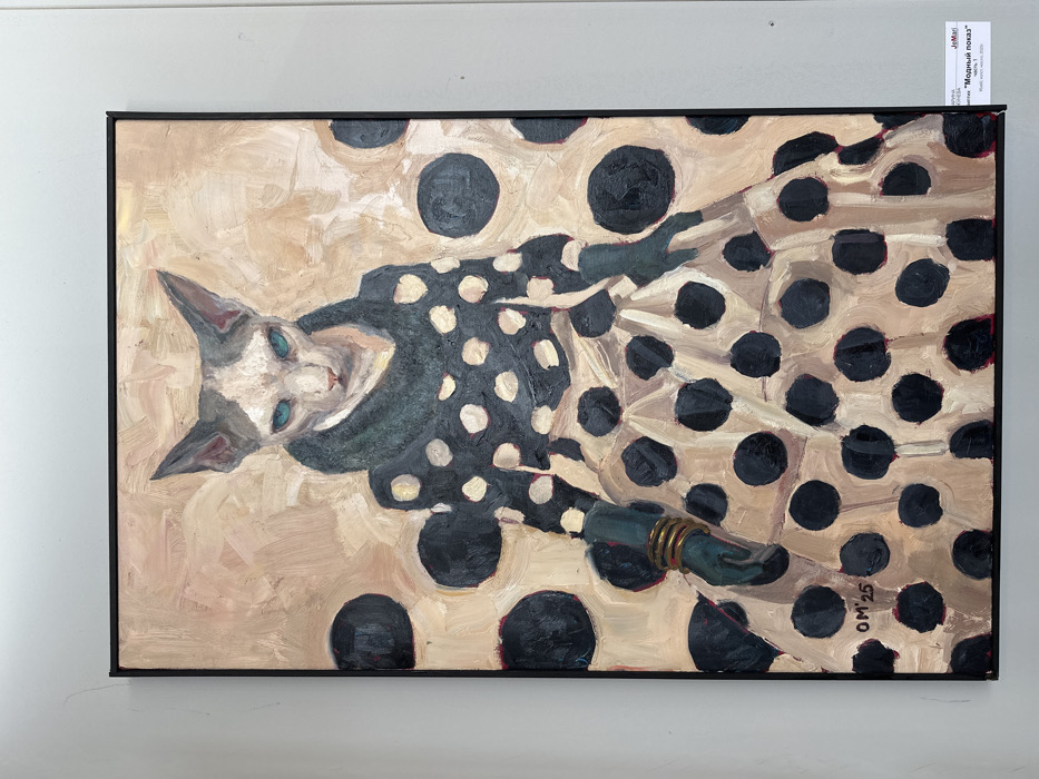 An oil painting, framed in black, is displayed on a light gray wall in Nizhny Novgorod, Russia. The artwork presents a stylized, full-body depiction of a Sphynx cat, rendered with visible brushstrokes. The cat faces forward, exhibiting a pale complexion with gray and white markings on its head and prominent turquoise eyes. It is dressed in human-like attire, featuring a dark teal turtleneck, a long-sleeved top patterned with white polka dots on a black background, and a voluminous skirt in light beige with large black polka dots. Dark teal gloves cover its hands, with multiple gold-colored bangles visible on its left wrist. The painting's background is a textured light beige, accented with scattered large black polka dots that echo the skirt's design. The initials "OM" and the number "25" are inscribed in the lower left corner of the canvas. Adjacent to the bottom right of the frame, a white label indicates the artwork's title as "Модный показ (Fashion Show) часть 1," attributed to artist "Ирина АНДРЕЕВА (Irina Andreeva)," and notes its medium as "Холст, масло (Canvas, oil)," dimensions as "80х50см," and production year as "2024."