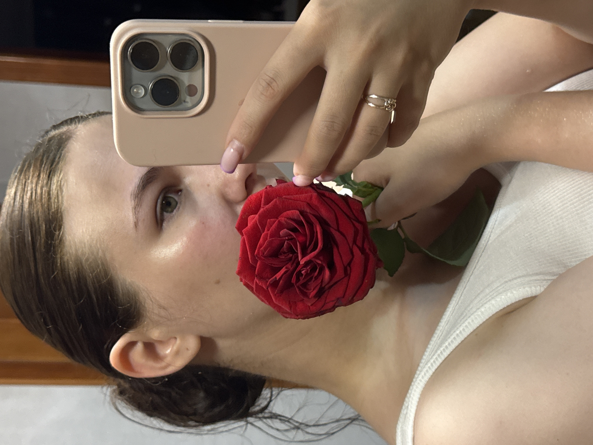 A young woman is taking a selfie in a mirror, holding a red rose to her face. She is wearing a white tank top. The background appears to be a room with wooden doors and a plain wall. The overall lighting suggests an indoor setting, possibly in a residential area of Chaikovsky, Russia. The woman's expression is partially obscured by the rose and the phone. The image captures a moment of personal reflection and self-expression.