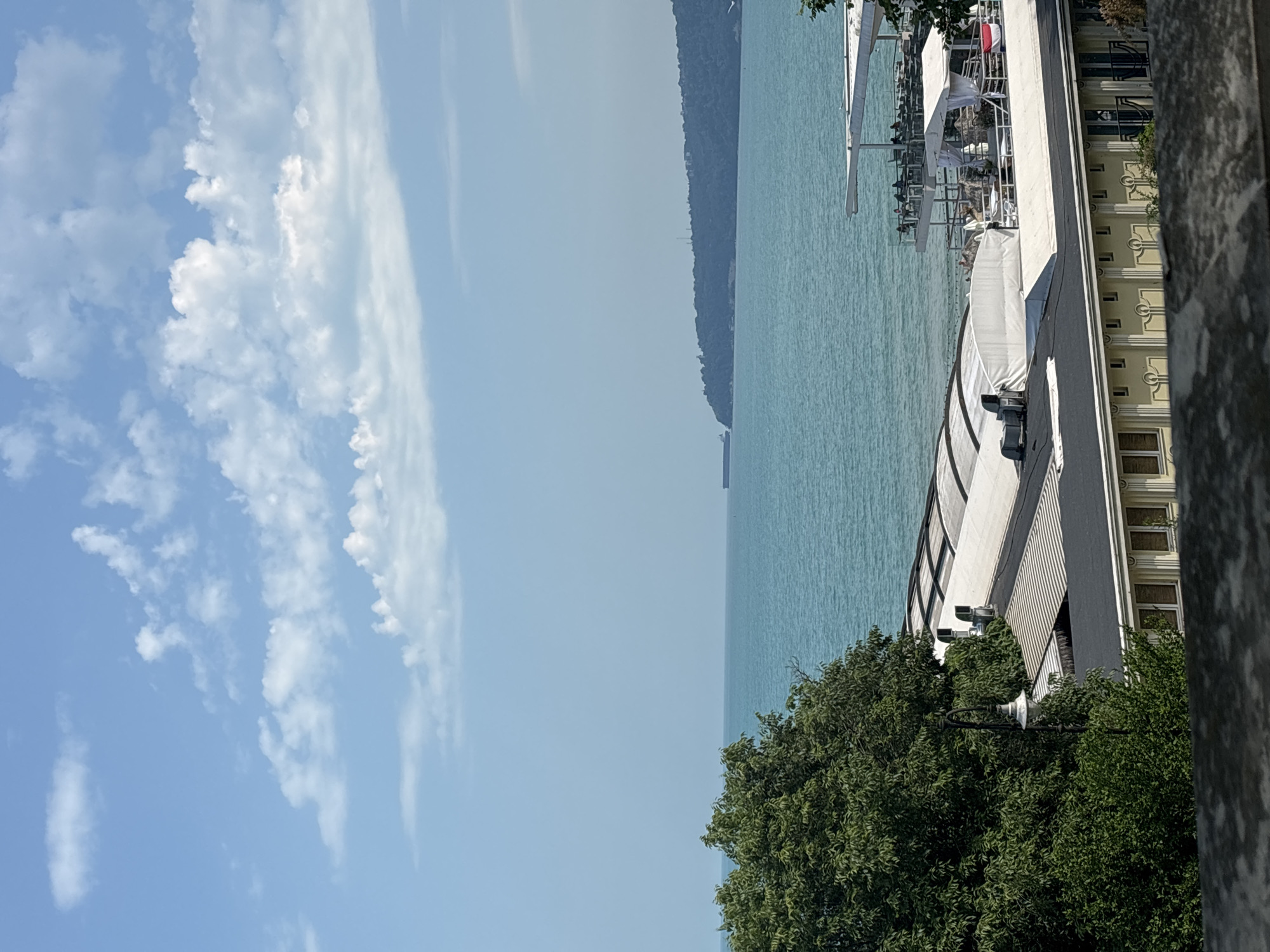 The image displays a serene coastal landscape under a bright, clear blue sky, dotted with scattered white clouds, some fluffy and some wispy. A vast expanse of calm, turquoise-blue water, consistent with the Black Sea, dominates the middle ground. In the distance, a dark green, forested landmass or hill forms the horizon, separating the water from the sky.
On the right side of the frame, a light-colored building, possibly a hotel or apartment complex, is prominent, featuring multiple windows and architectural details. Below it, a white structure, likely a terrace, restaurant, or pier, extends towards the water, covered by white awnings and umbrellas, suggesting outdoor seating. Lush green trees in the foreground partially obscure these structures and the water, adding natural texture to the scene. A grey concrete or stone pillar frames the extreme right edge of the image.
The scene captures a peaceful and sunny day, likely mid-morning or early afternoon, indicated by the strong illumination. The absence of specific human activity suggests a tranquil atmosphere, but the presence of leisure-oriented structures implies potential for relaxation and dining by the sea. The overall setting, with its distinctive turquoise water and coastal architecture, aligns with a resort location like Varna, Bulgaria. No visible text is present in the image.