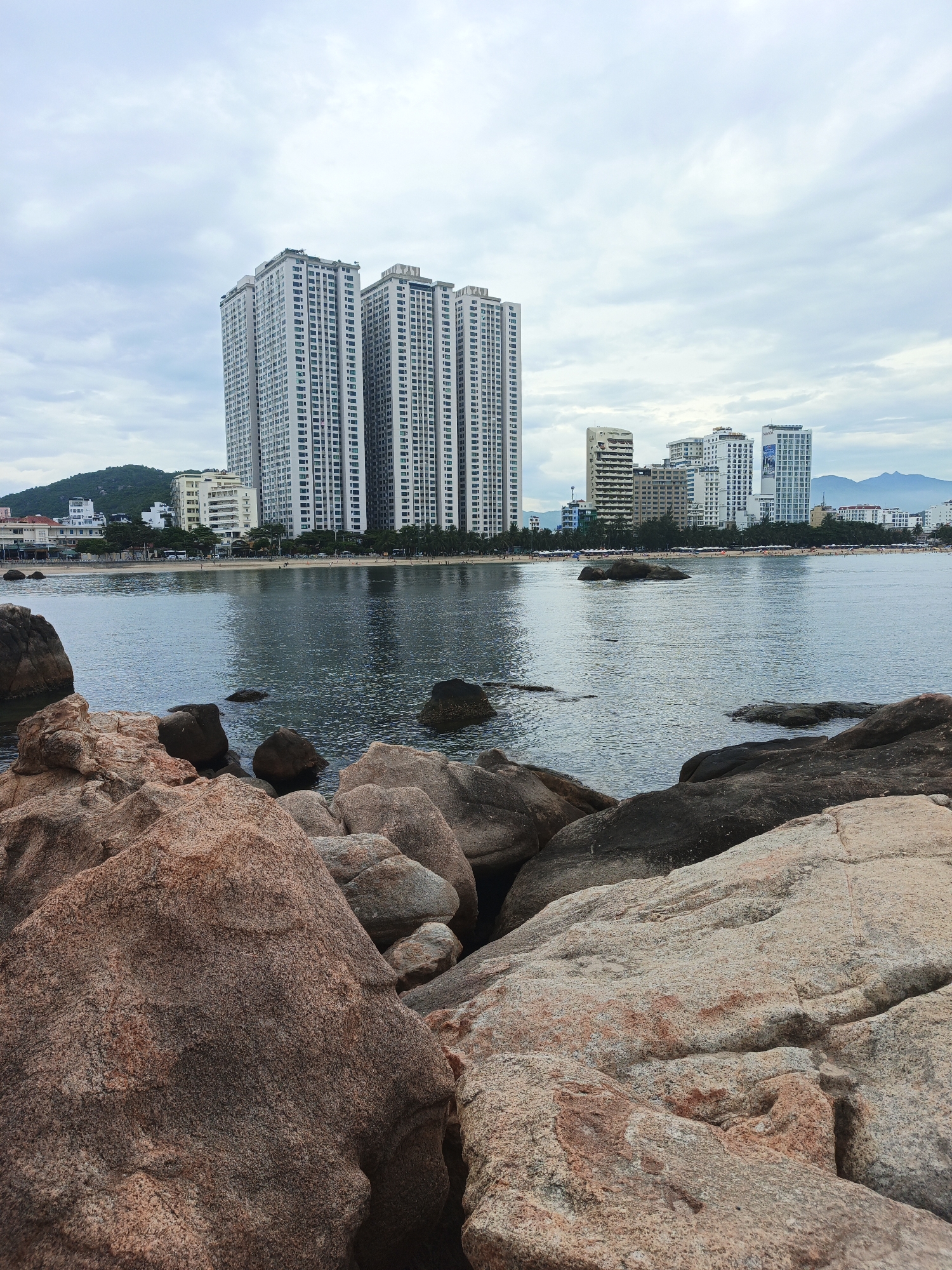 The image depicts a scenic view of the coastline in Nha Trang, Vietnam. In the foreground, large, weathered rocks are scattered along the shore, with some partially submerged in the calm, dark water. Beyond the rocks, a sandy beach stretches out, lined with palm trees and various buildings. Dominating the skyline are several tall, modern apartment buildings and hotels, characteristic of Nha Trang's urban development. The sky is overcast with grey clouds, suggesting a cloudy day. The water appears tranquil, with gentle ripples on its surface. The overall scene captures the blend of natural beauty and urban architecture that defines the Nha Trang waterfront.