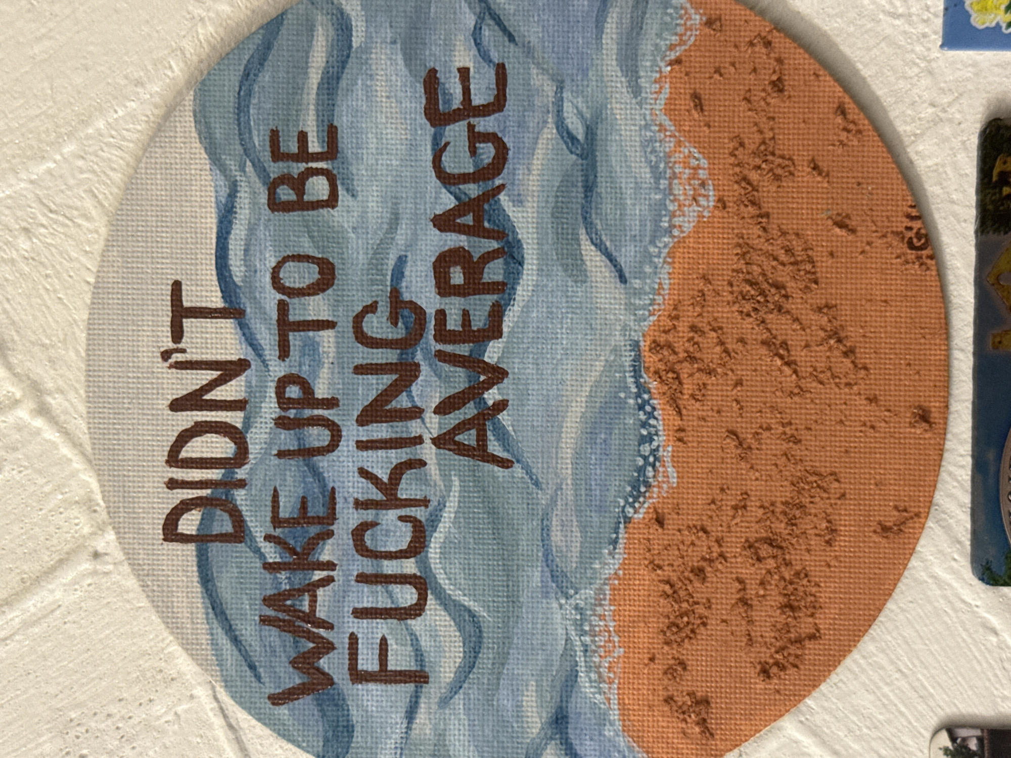 A circular canvas painting depicts a beach scene with the inscription "DIDN'T WAKE UP TO BE FUCKING AVERAGE" in brown lettering. The left half of the canvas shows blue and white waves, while the right half depicts a tan sandy beach. The painting is attached to a textured, white surface. Other objects including a picture of yellow flower and a park sign are visible in the background. The location in Asenovgrad, Bulgaria is not determinable from this image.