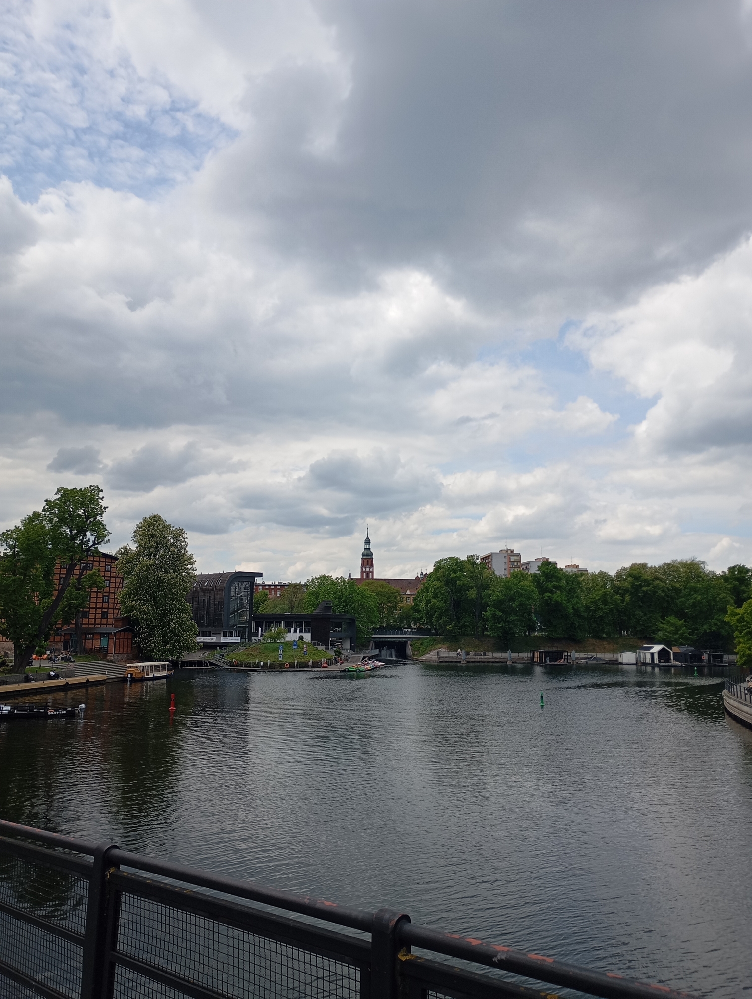 This photograph depicts a view of the Brda River in Bydgoszcz, Poland. The foreground contains a dark railing with a wire mesh infill. Behind the railing is the Brda River, reflecting the overcast sky. Across the river, the scene includes a mix of buildings, trees, and waterfront structures. Buildings appear to be of varying architectural styles, with one in the center-right bearing a distinctive tower. There is a small island with a restaurant and seating area. The sky is predominantly cloudy, with some patches of blue visible. The water is calm, with gentle ripples, and various small boats and structures line the banks. No individuals are clearly visible.
