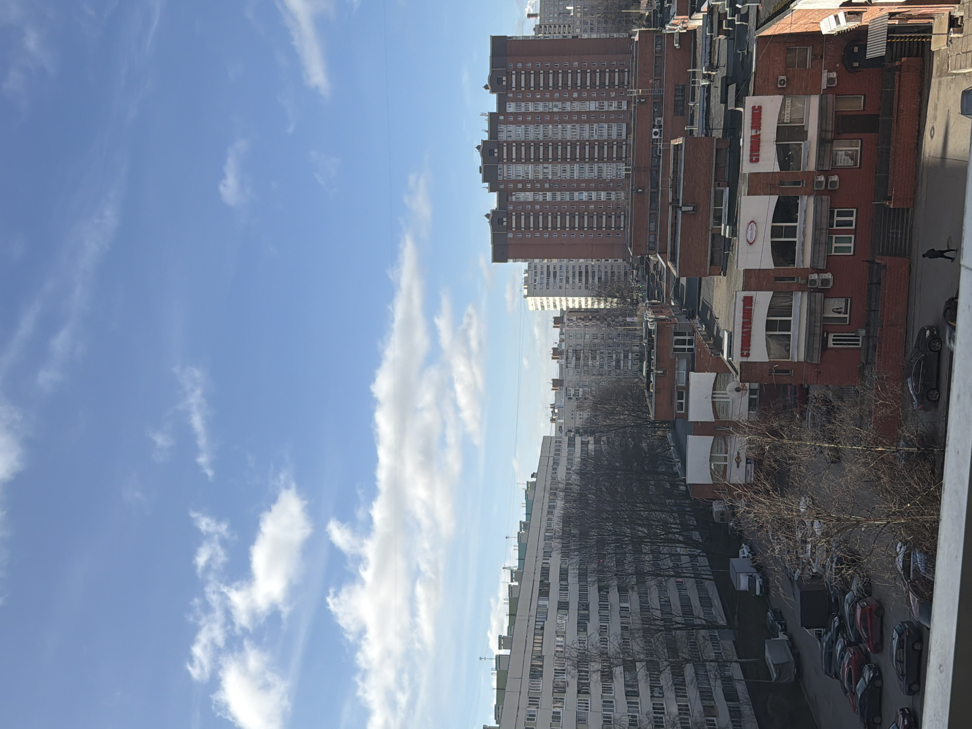 The image is a medium-angle outdoor shot depicting a cityscape under a blue sky with scattered clouds. The scene appears to be Saint Petersburg, Russia, identified by the architectural style of the buildings. The right side of the image features red brick buildings of varying heights, some displaying signage written in Cyrillic, possibly indicating commercial or residential establishments. Cars line the street and a pedestrian walks on the sidewalk. On the left, taller, white buildings with numerous windows stretch into the distance. Bare trees without leaves suggest it is either late autumn or early spring.